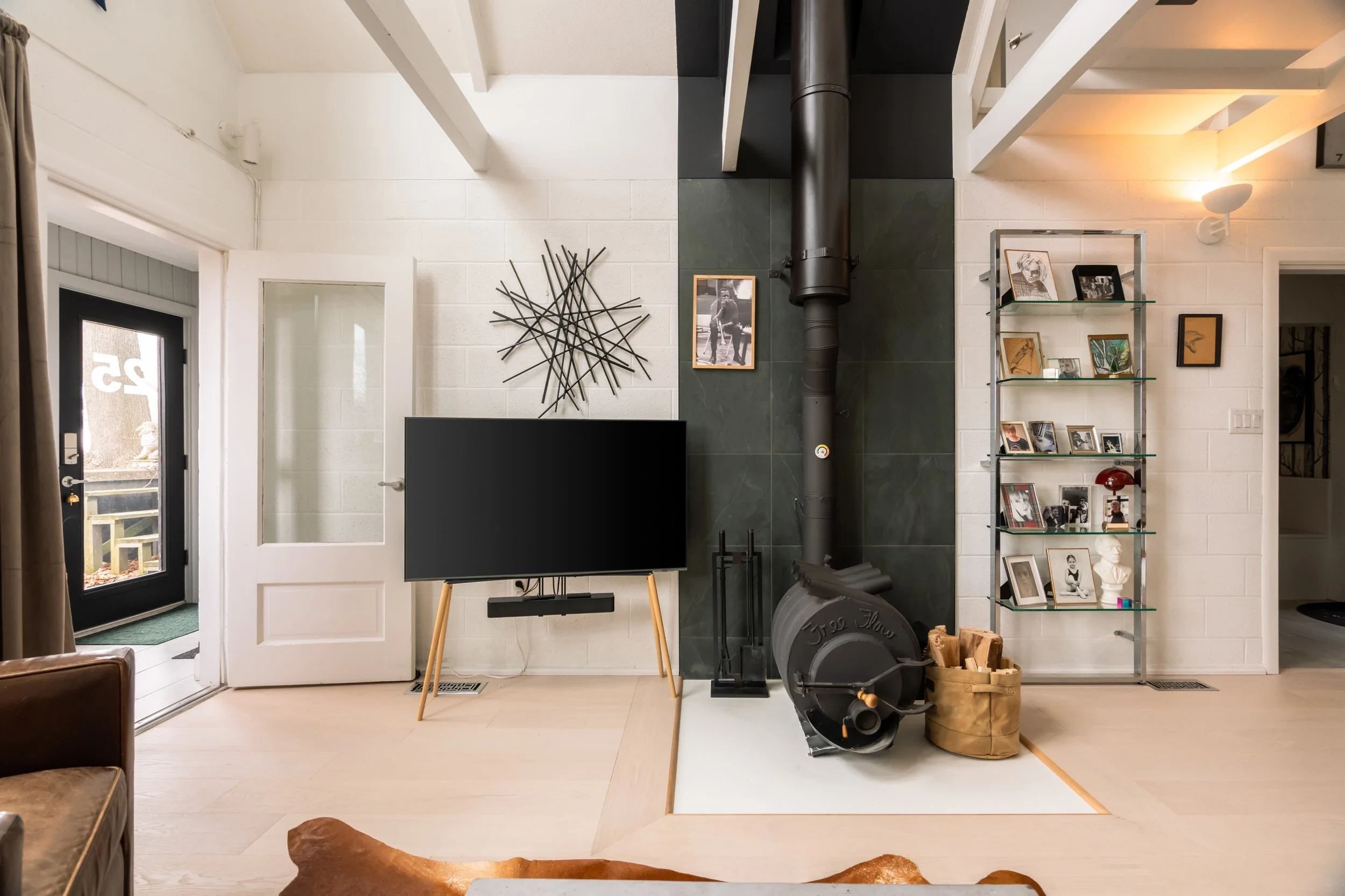 Living room with a door leading outside, a black television on a wooden stand, a decorative wall art piece, a framed photo on the wall, a bookshelf with photos and decorations, and a black wood stove on a white hearth.