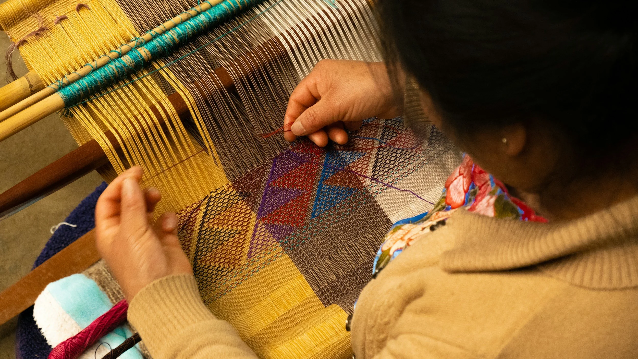 A person weaving a colorful, geometric patterned textile on a loom with yellow, purple, blue, and red threads.