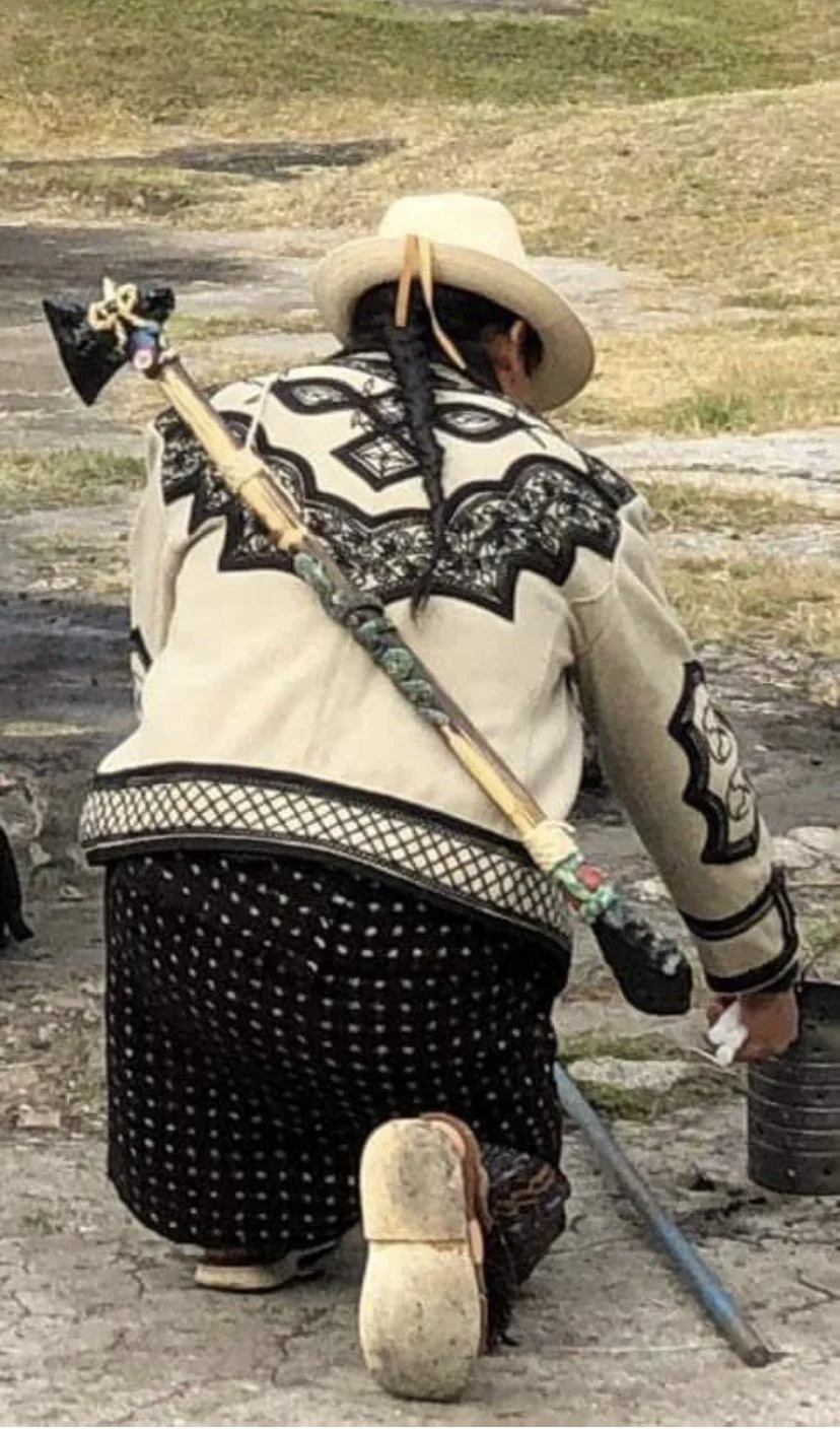 A woman dressed in traditional Mexican clothing, with a large woven hat, is seen squatting outdoors. She is carrying a decorated stick or staff over her shoulder and is holding a small container or bucket.
