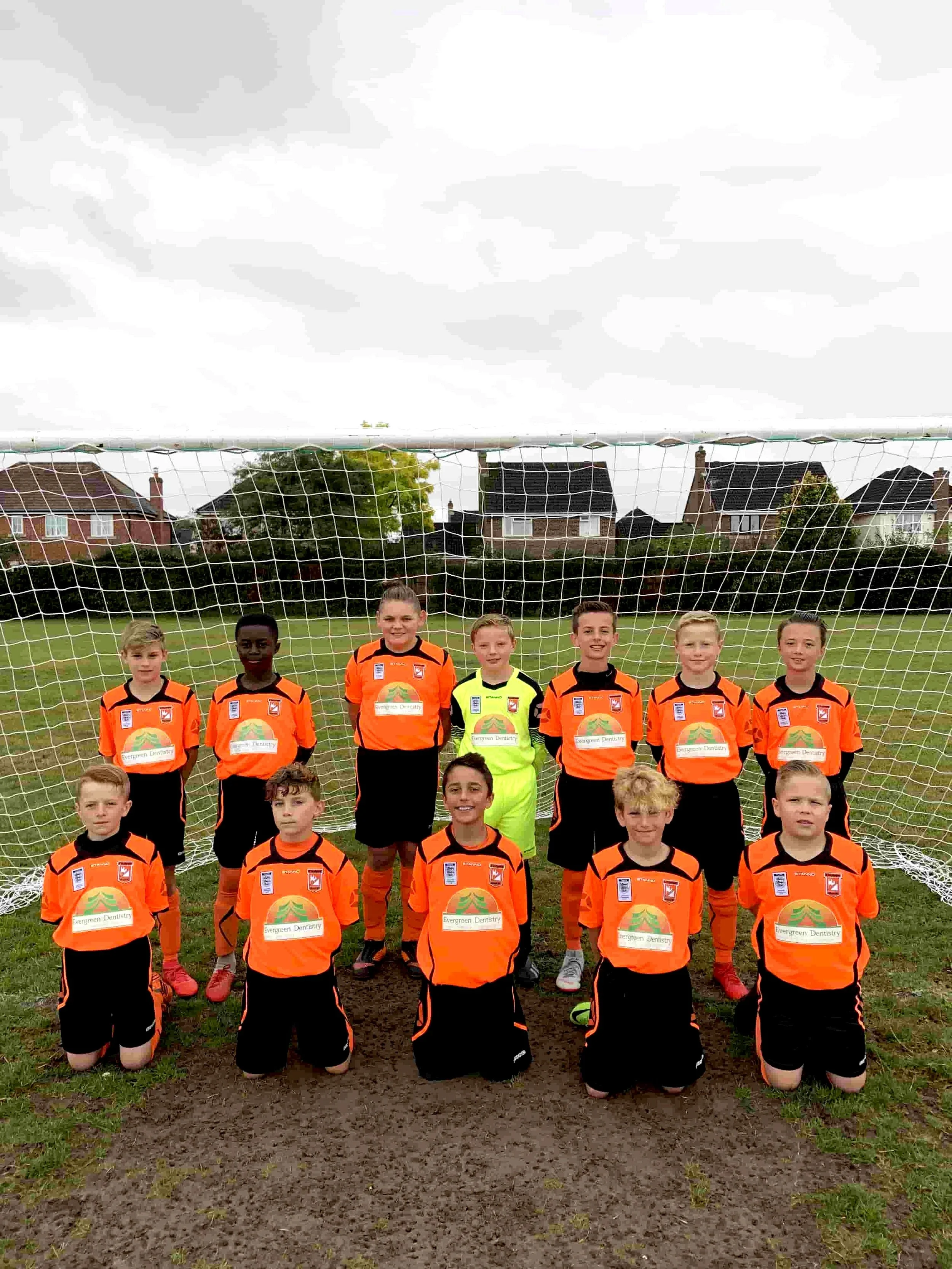 Youth soccer team in orange uniforms posing in front of a goal on a grassy field.