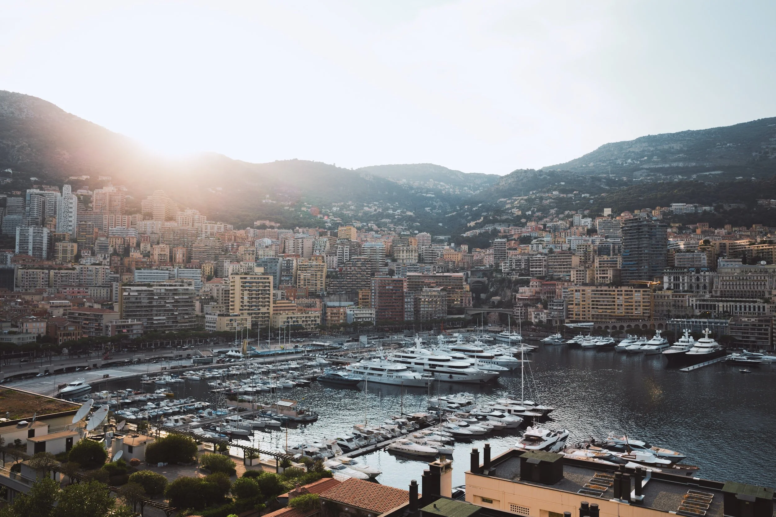 City skyline with numerous high-rise buildings and a marina filled with yachts and boats, set against mountainous terrain with the sun shining from behind the mountains.
