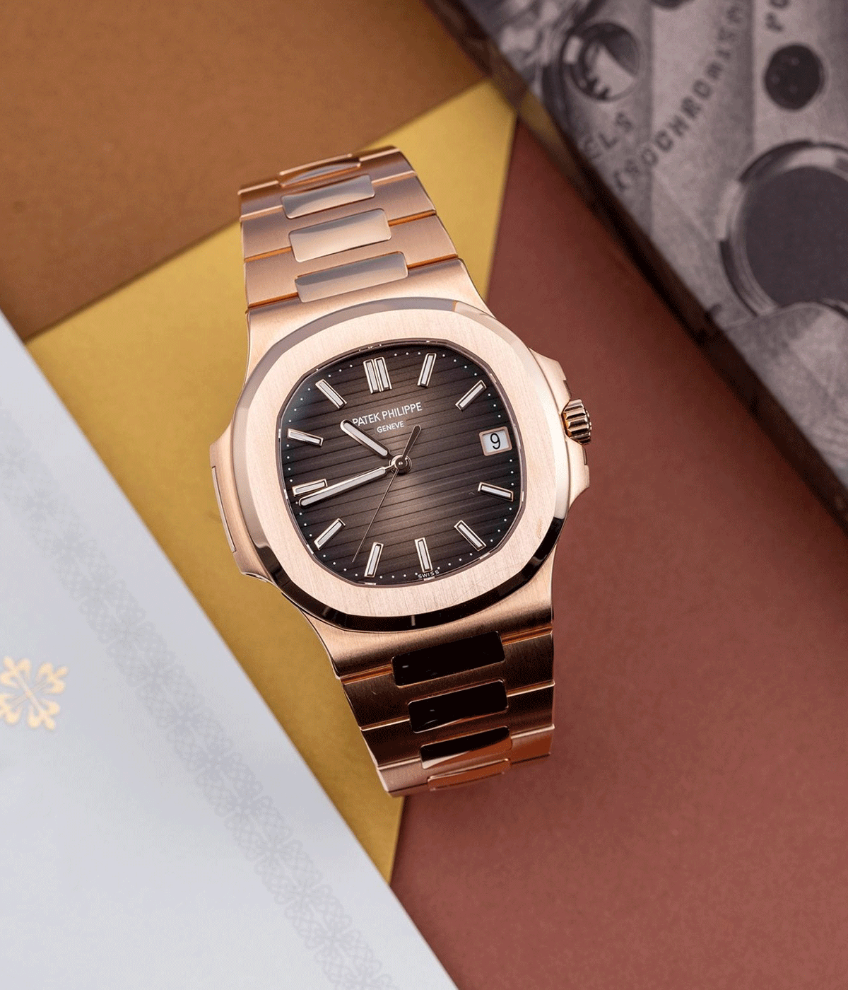 A rose gold Patek Philippe wristwatch with a black textured dial, Roman numeral markers, and a date window at 3 o'clock, resting on colored geometric paper backgrounds.