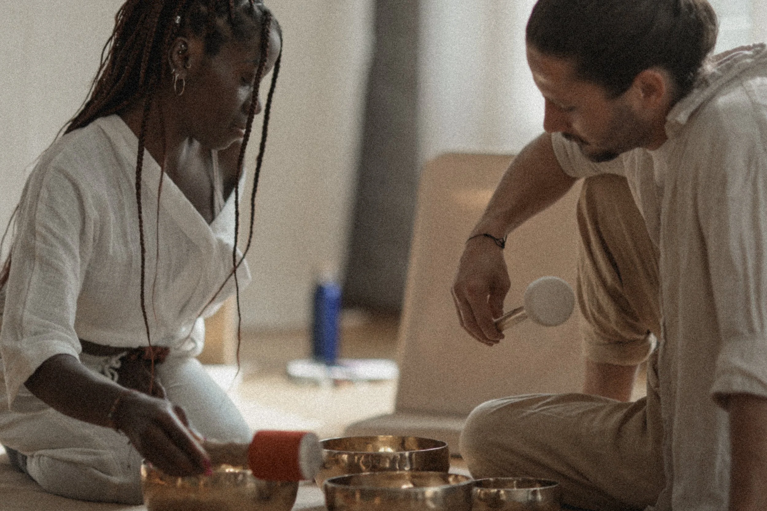 A woman and a man sit cross-legged on the floor, engaging in a spiritual or ritual practice with singing bowls, in a softly lit room.