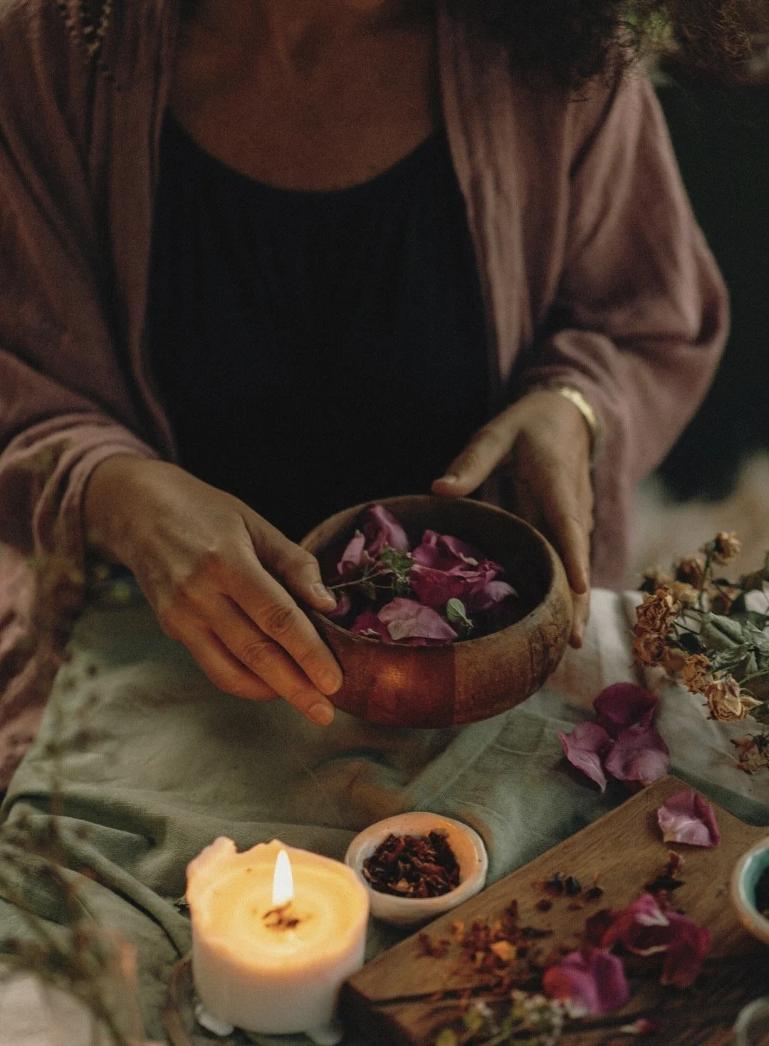 A person holding a wooden bowl filled with pink flower petals, with a lit candle, dried herbs, and scattered petals on the table.