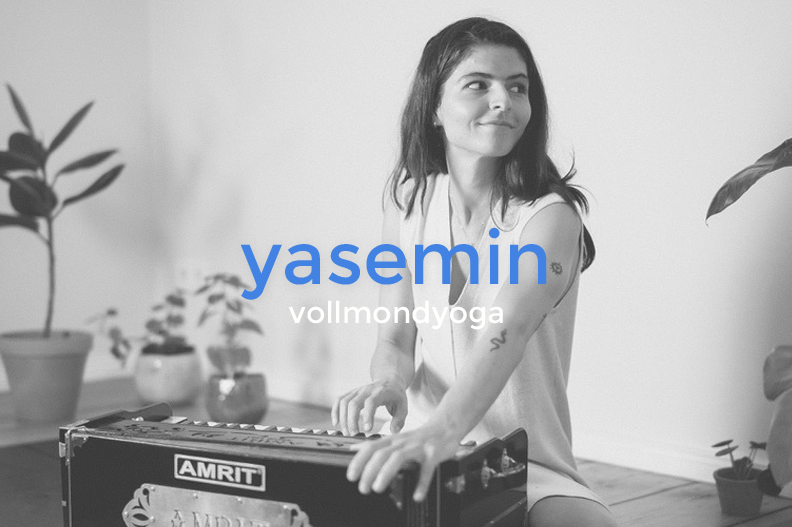 A young woman with dark hair sits on the floor, playing a harmonium, surrounded by potted plants, with a relaxed smile on her face.