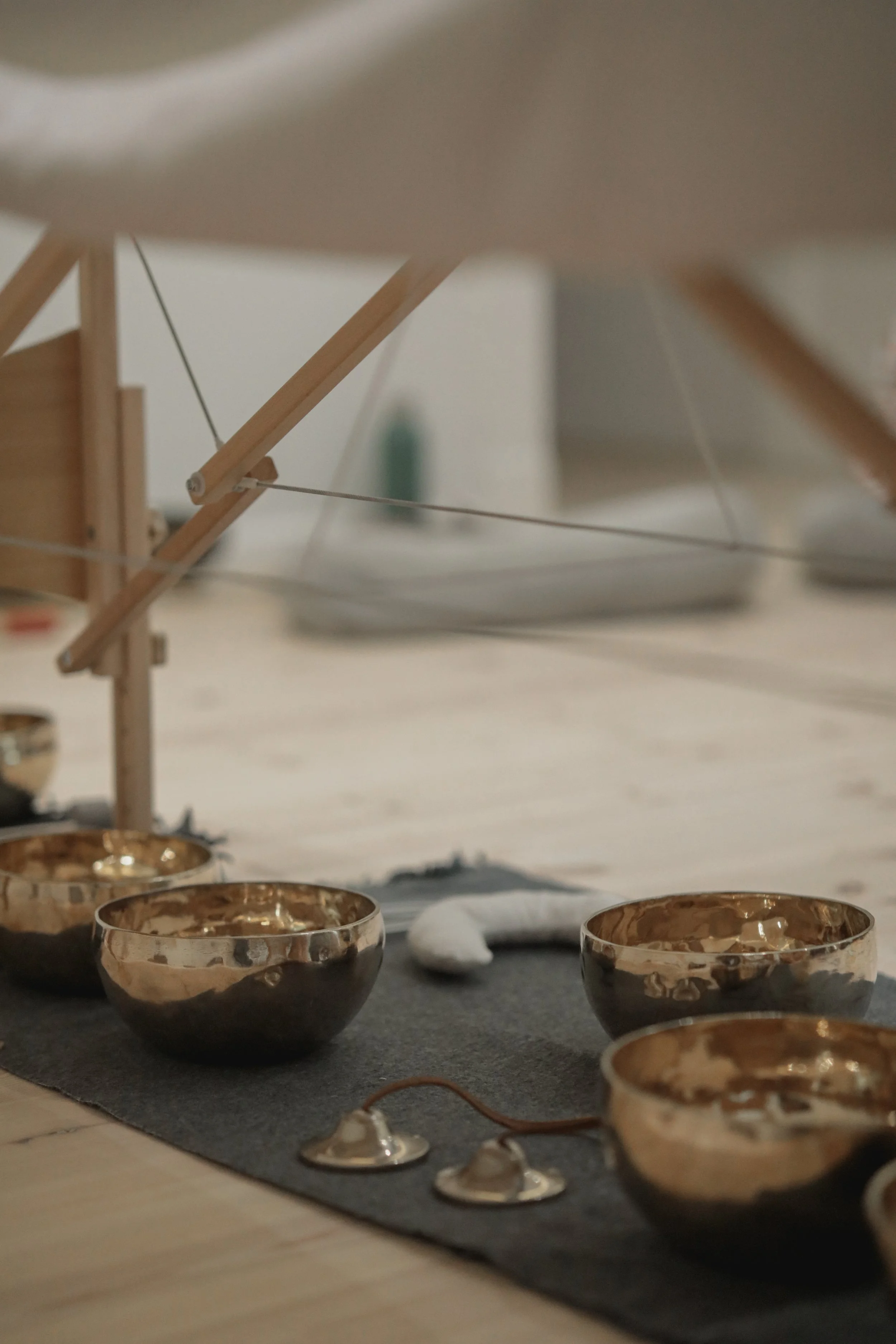 A set of copper singing bowls placed on a black mat with a wooden and string structure behind them, likely a wind chime or musical instrument, on a light-colored wooden surface.