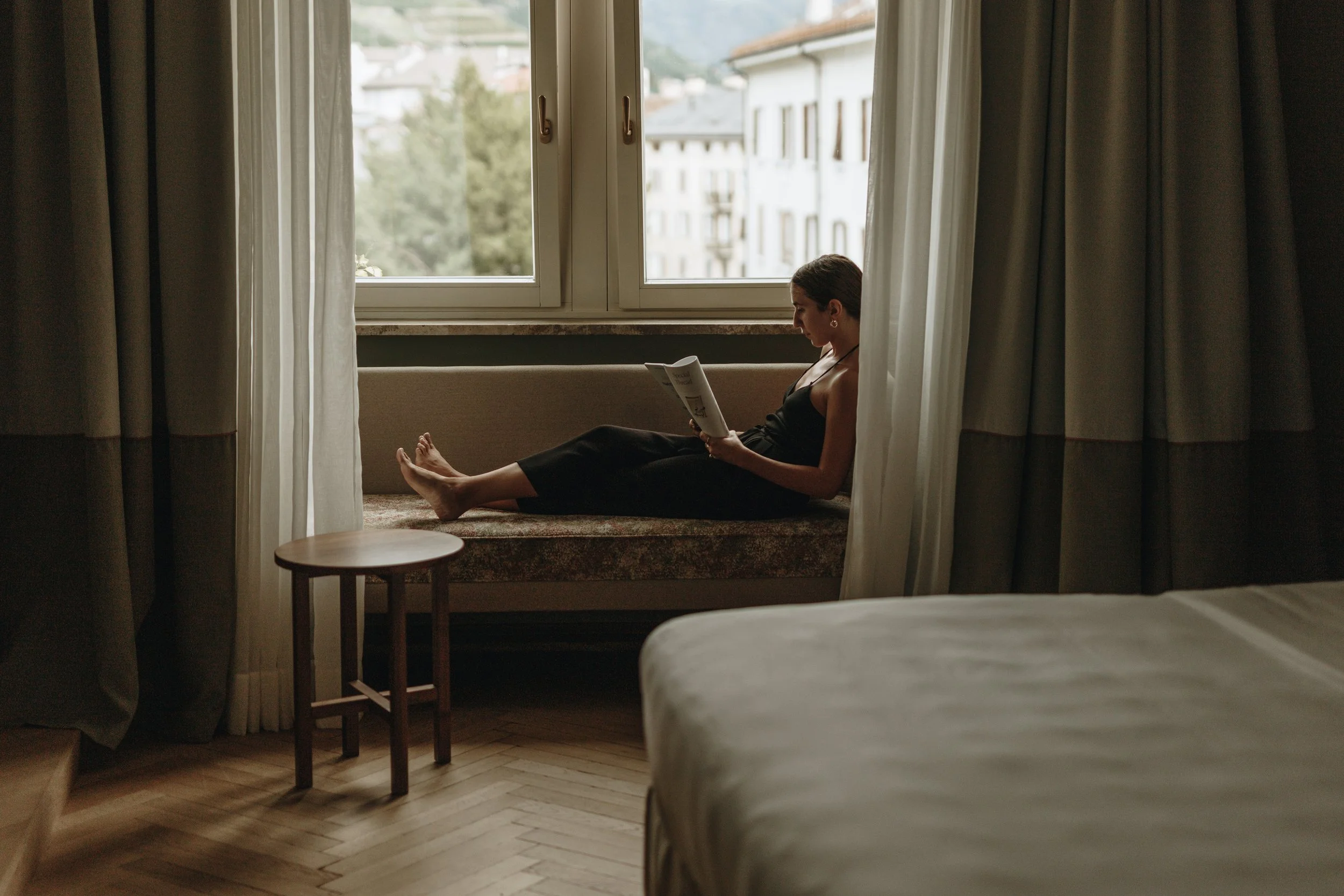 A woman is sitting on a window seat in a hotel room, reading a book or magazine. She is wearing a black sleeveless dress, with curtains on either side of the window and a small wooden table nearby.