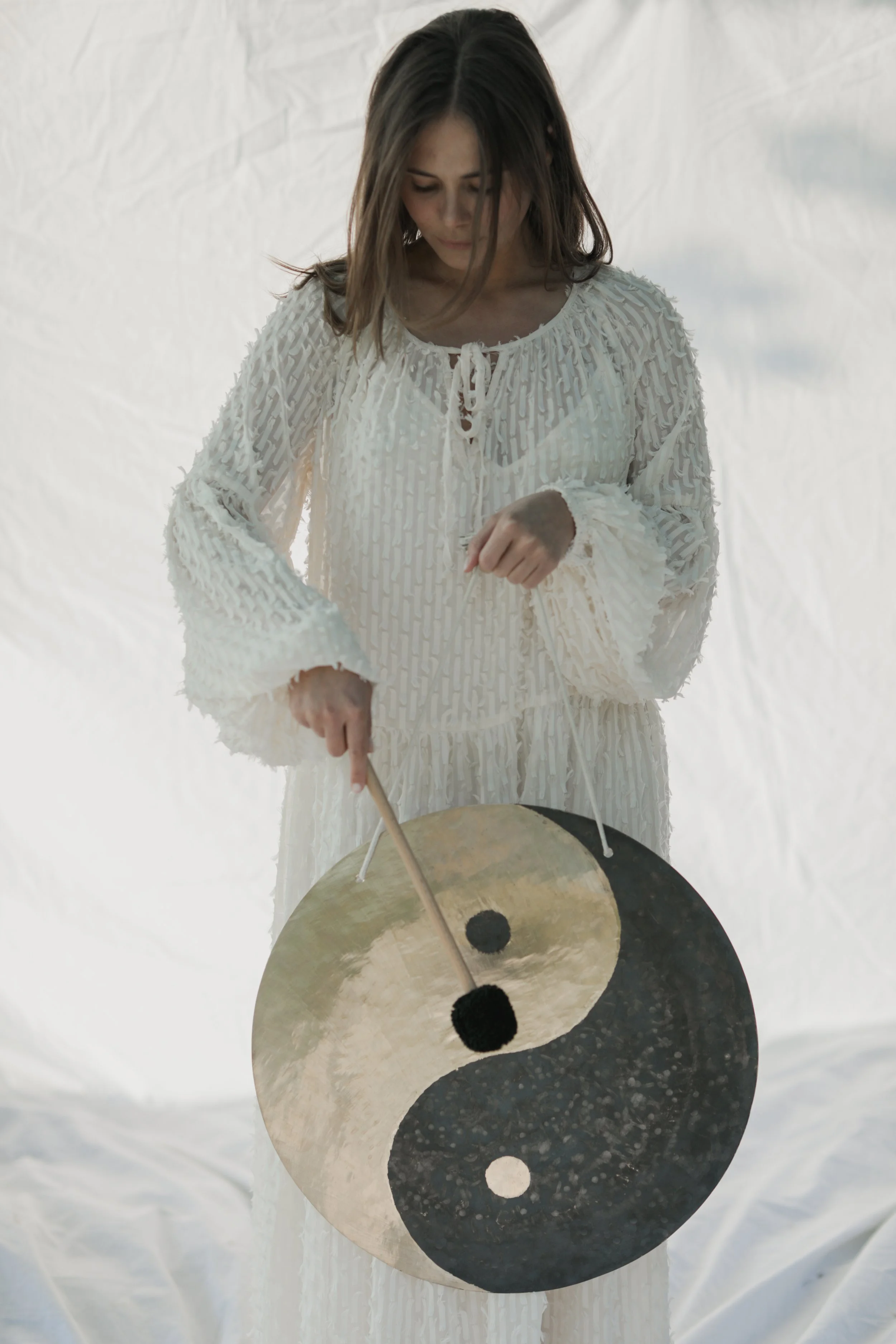 A woman in a white textured dress holds a yin-yang gong and a mallet, standing against a white background.