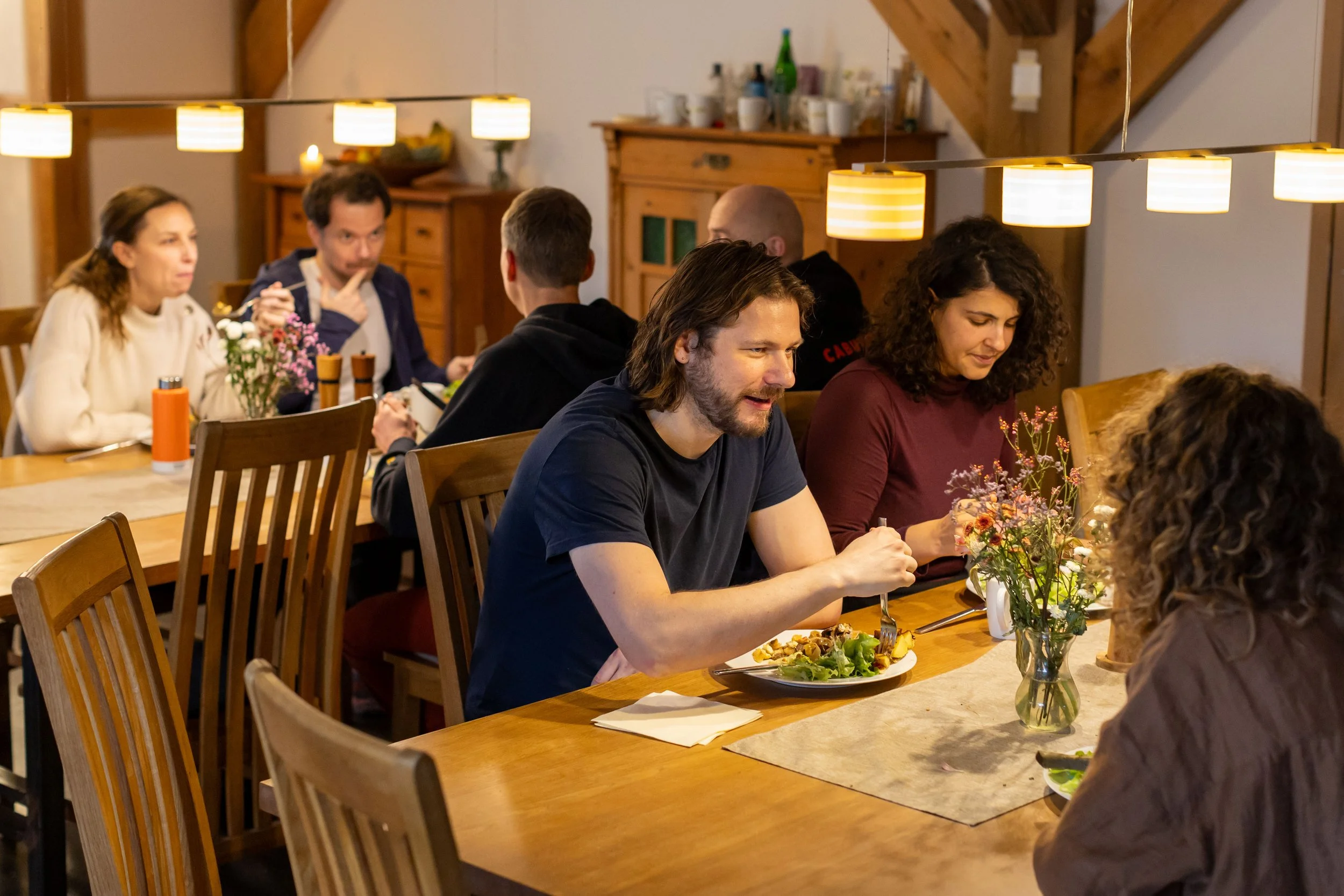 People sitting around a wooden dining table, eating and chatting in a cozy, well-lit room with flowers on the table.