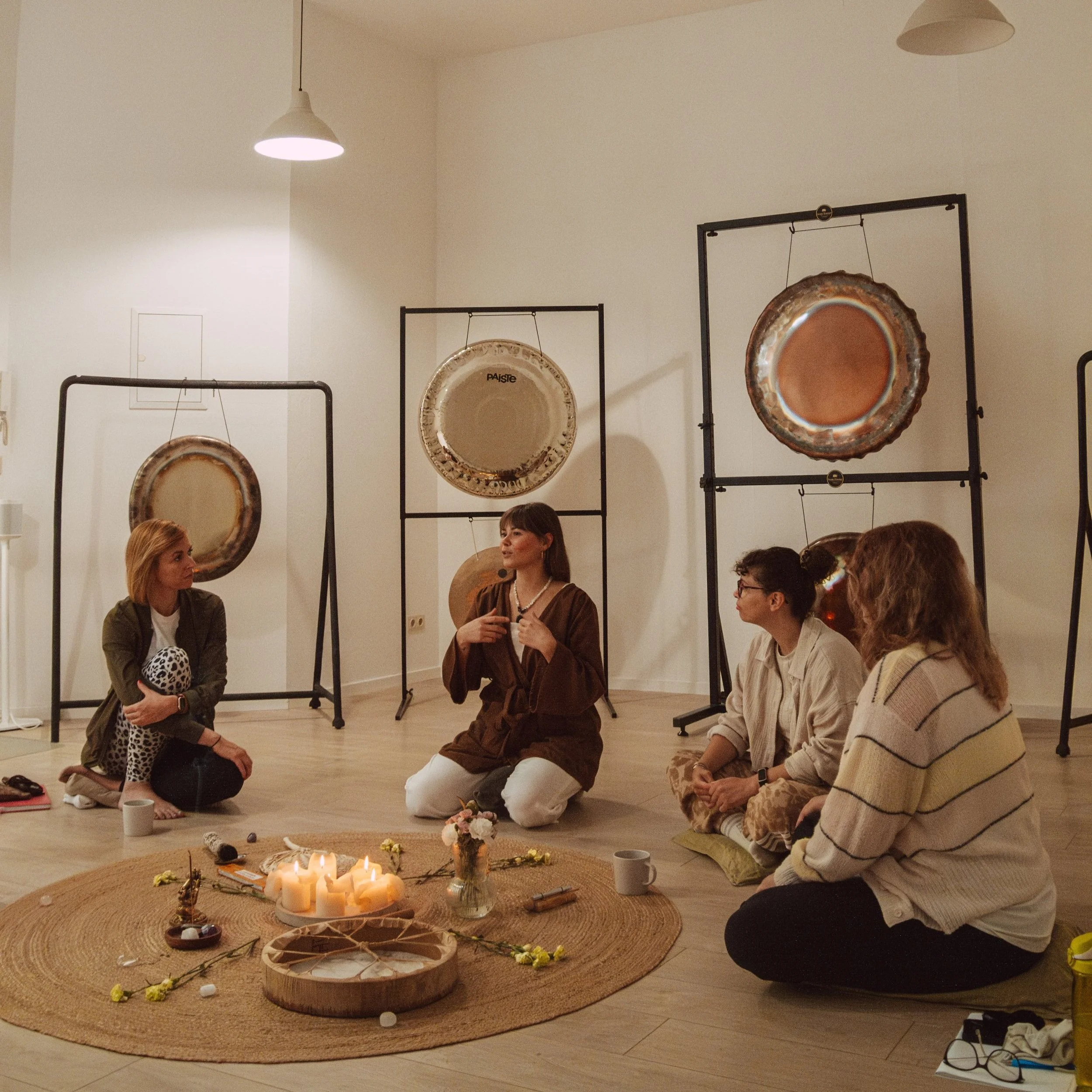 Four women sitting on the floor in a circle having a discussion in an art gallery with large metallic gong sculptures behind them. Candles and flowers are arranged on a circular woven mat in the center.