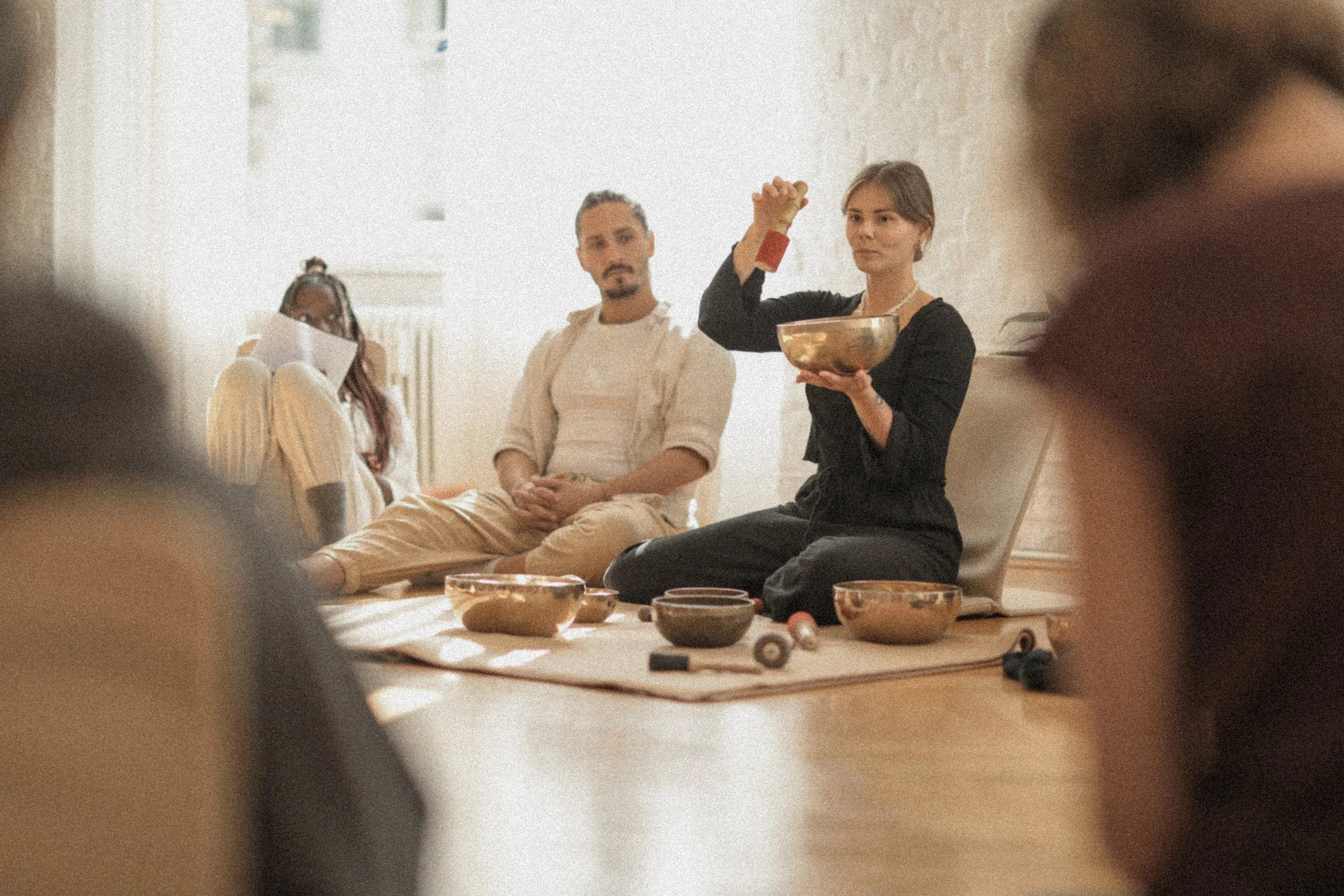 A woman demonstrating a spiritual or mystical practice with various bowls and tools on a mat, while others sit in a circle in a softly lit room.