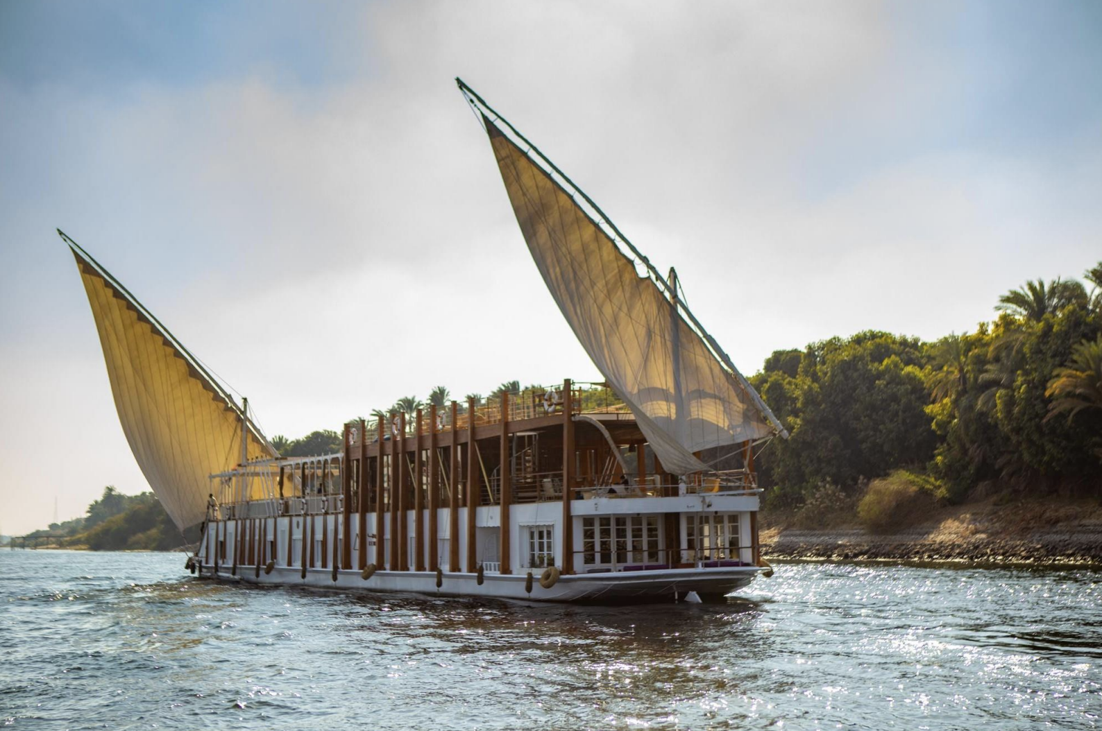 A boat with traditional sails sailing on a river near a lush, green shoreline.