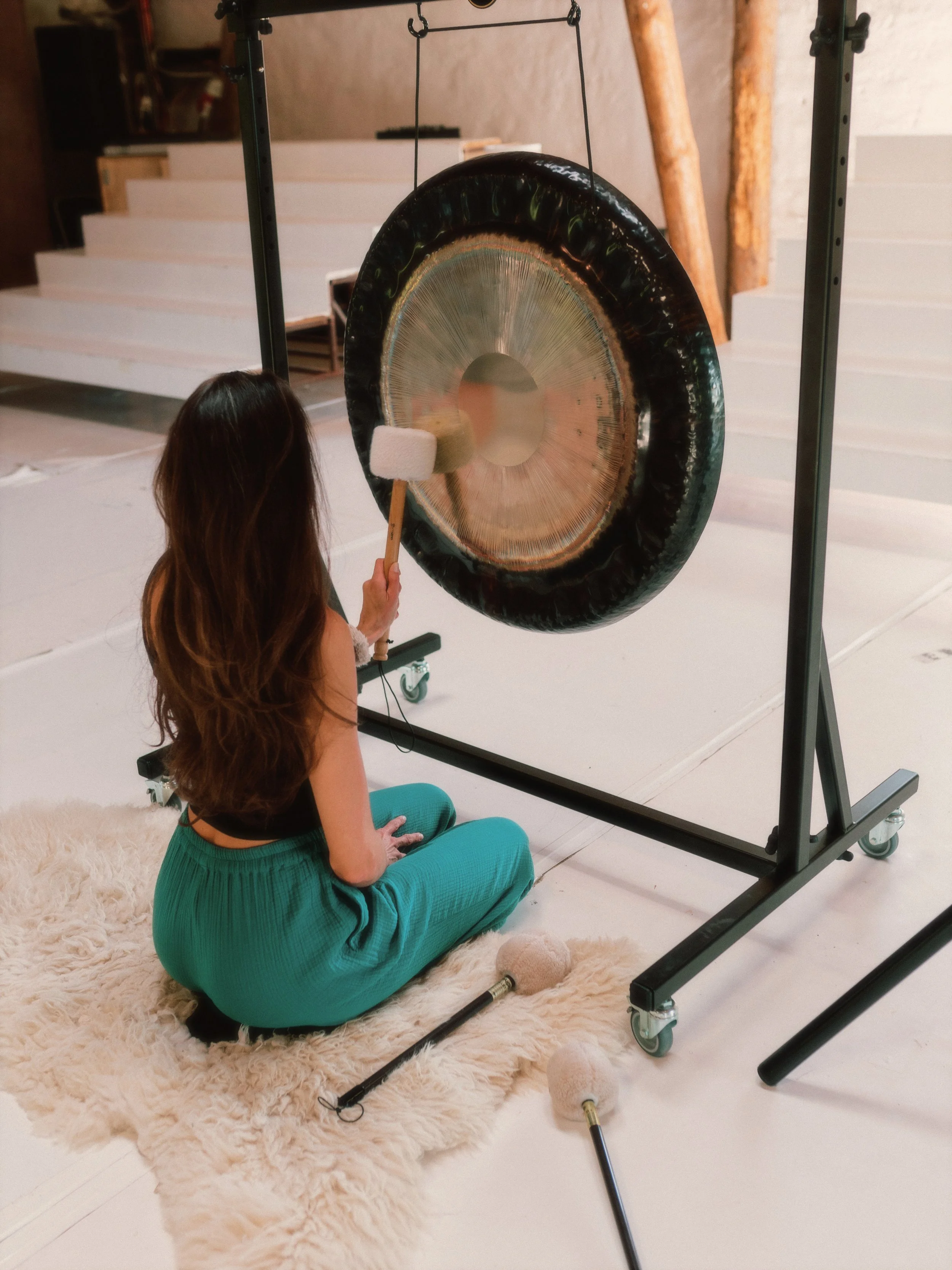 A woman with long brown hair, wearing teal pants and a black top, sitting cross-legged on a white furry rug, is hitting a large gong with a mallet. The gong hangs from a black metal frame with wheels.