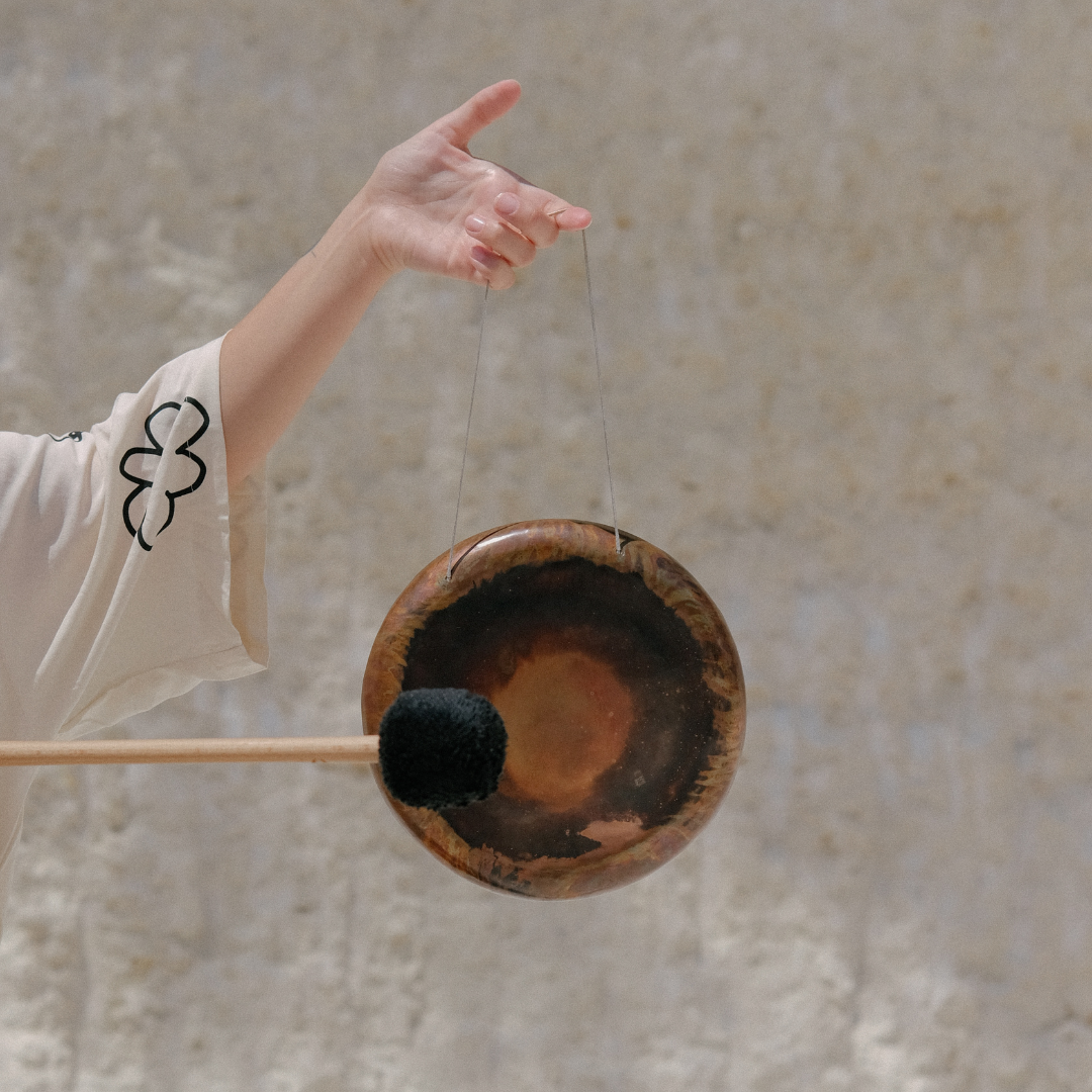 A person holding a wooden gong with a mallet, with a beige textured wall in the background.