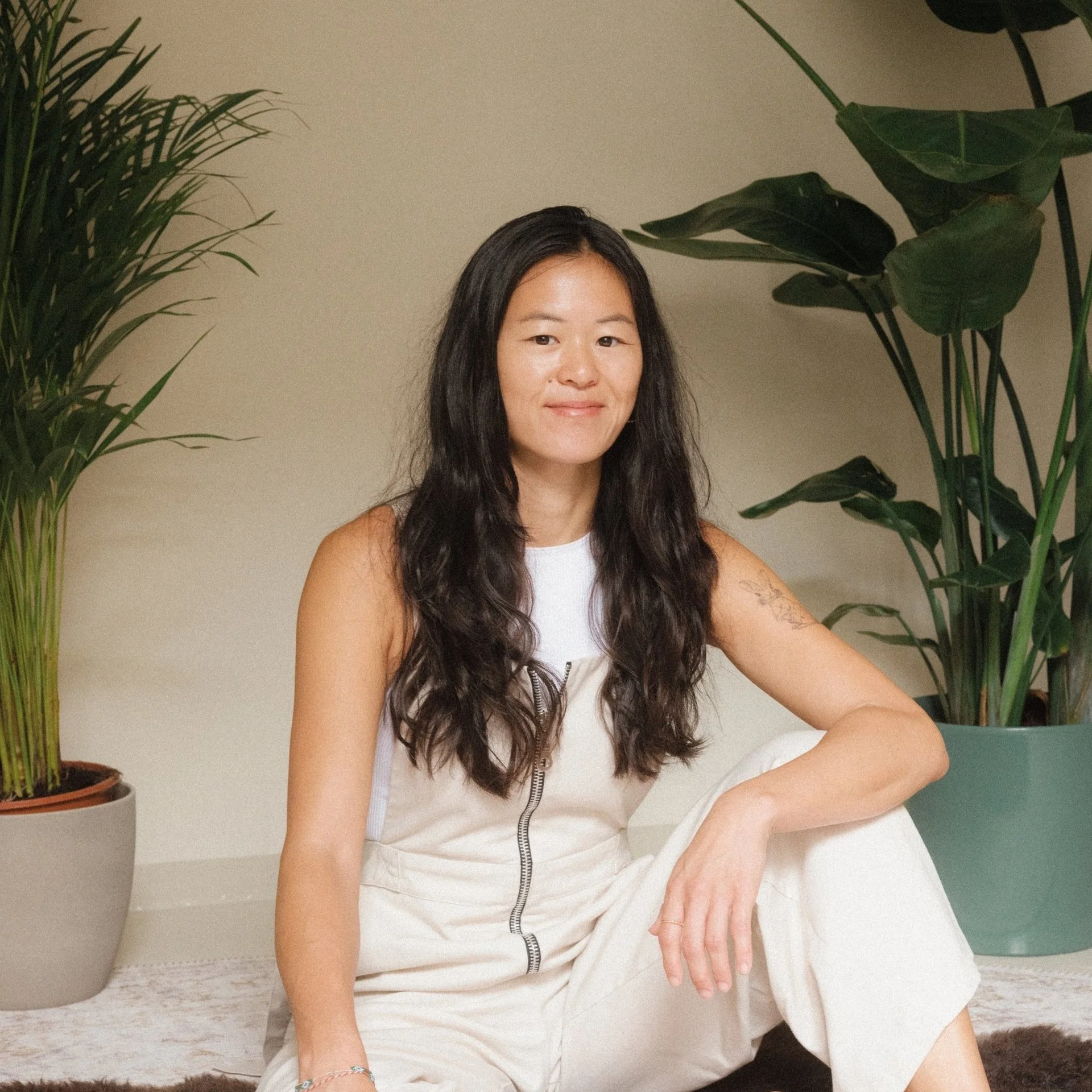 A woman with long dark wavy hair, wearing a sleeveless white top and cream-colored pants, sitting on the floor next to large potted green plants.