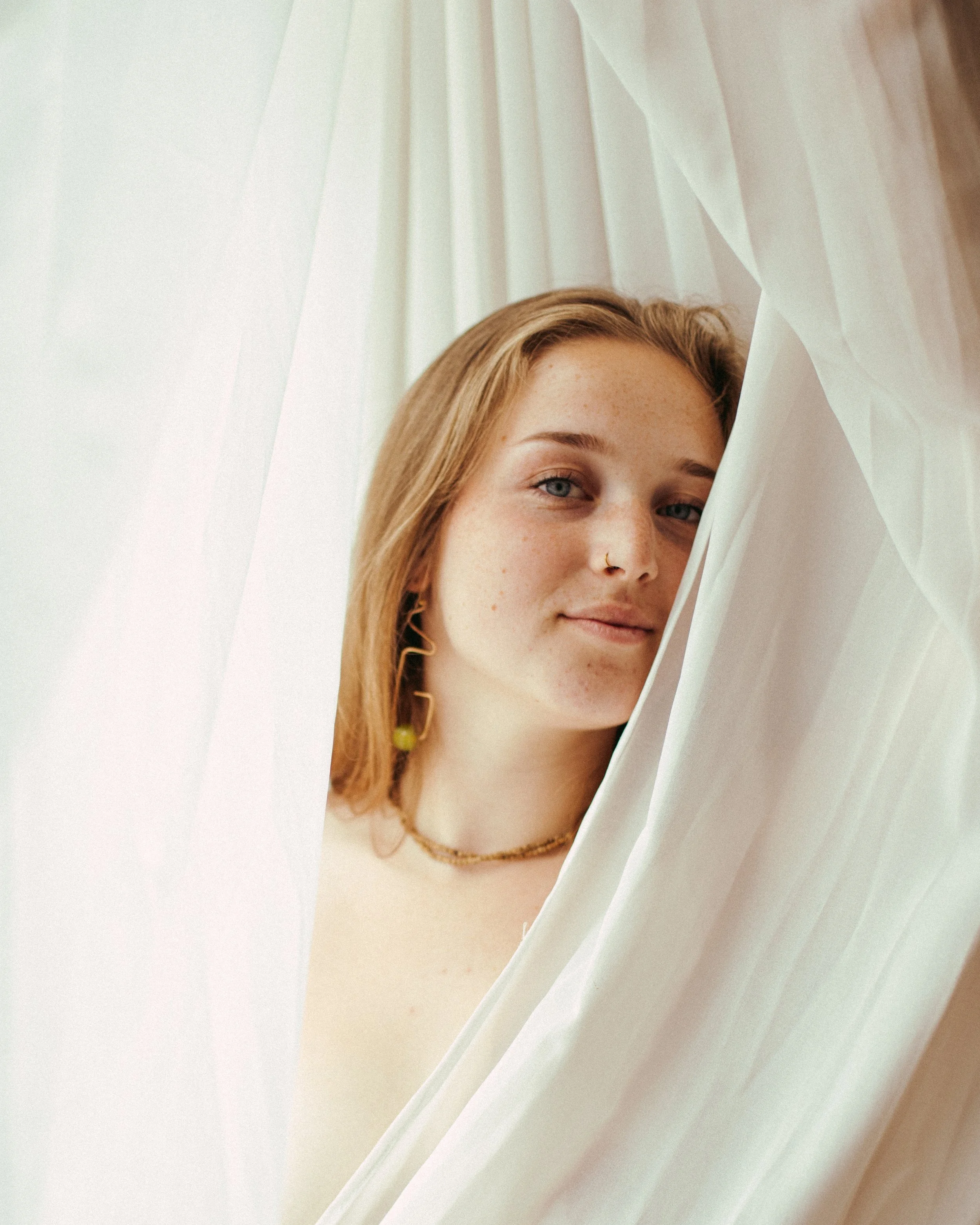 A young woman with fair skin, blonde hair, and blue eyes looks at the camera through a white curtain.
