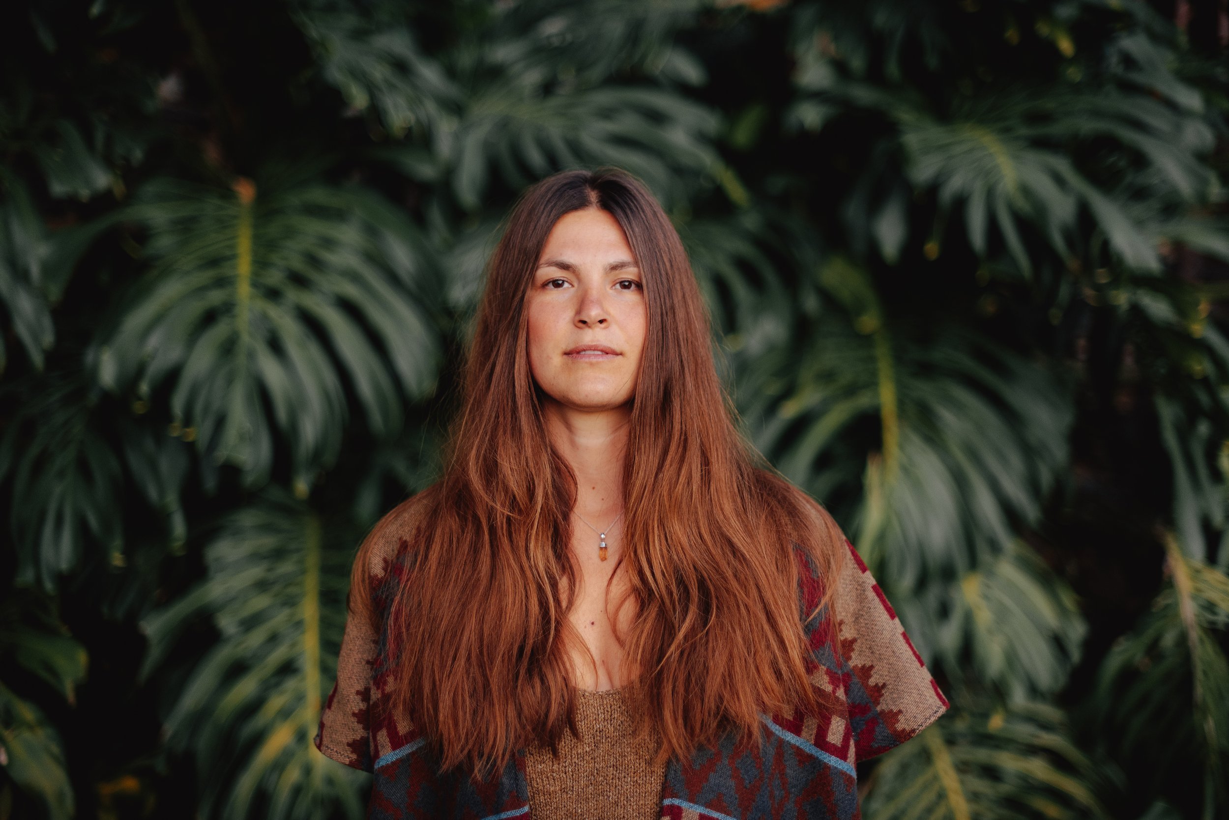 A woman with long, wavy reddish-brown hair, wearing a patterned jacket over a brown top, standing in front of lush green tropical plants.