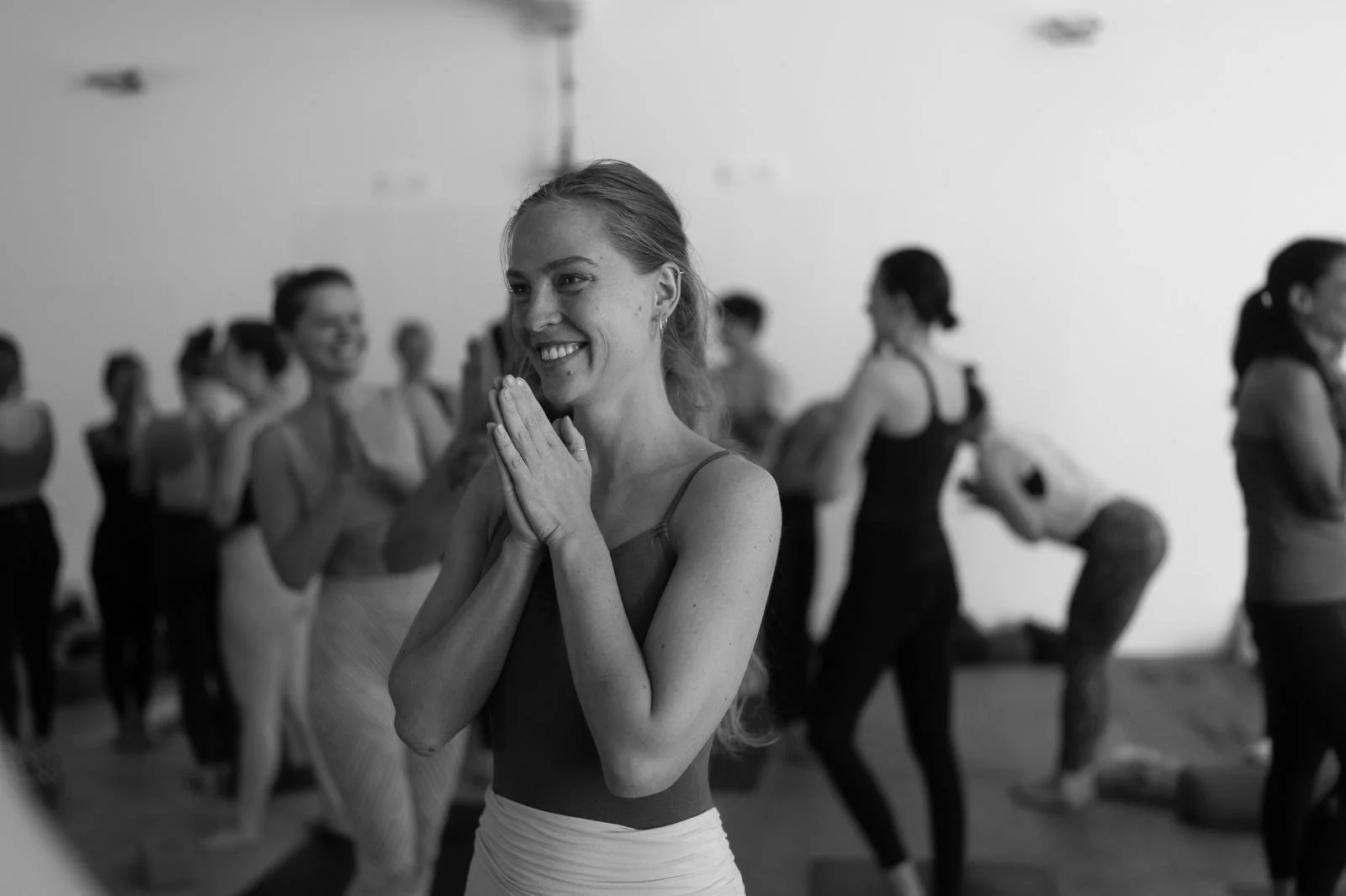 Young woman smiling with hands in prayer position in a dance or exercise studio, surrounded by other women, some practicing poses or stretches.
