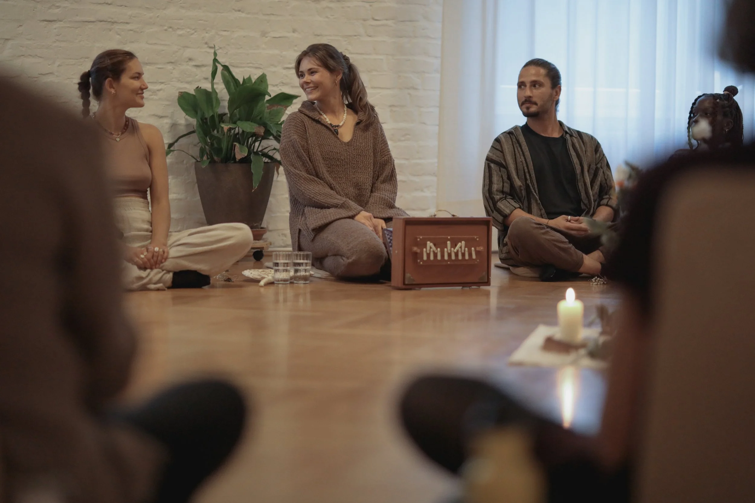 Four people sitting on the floor in a circle during a meditation or spiritual gathering, with a lit candle and a vintage wooden box.