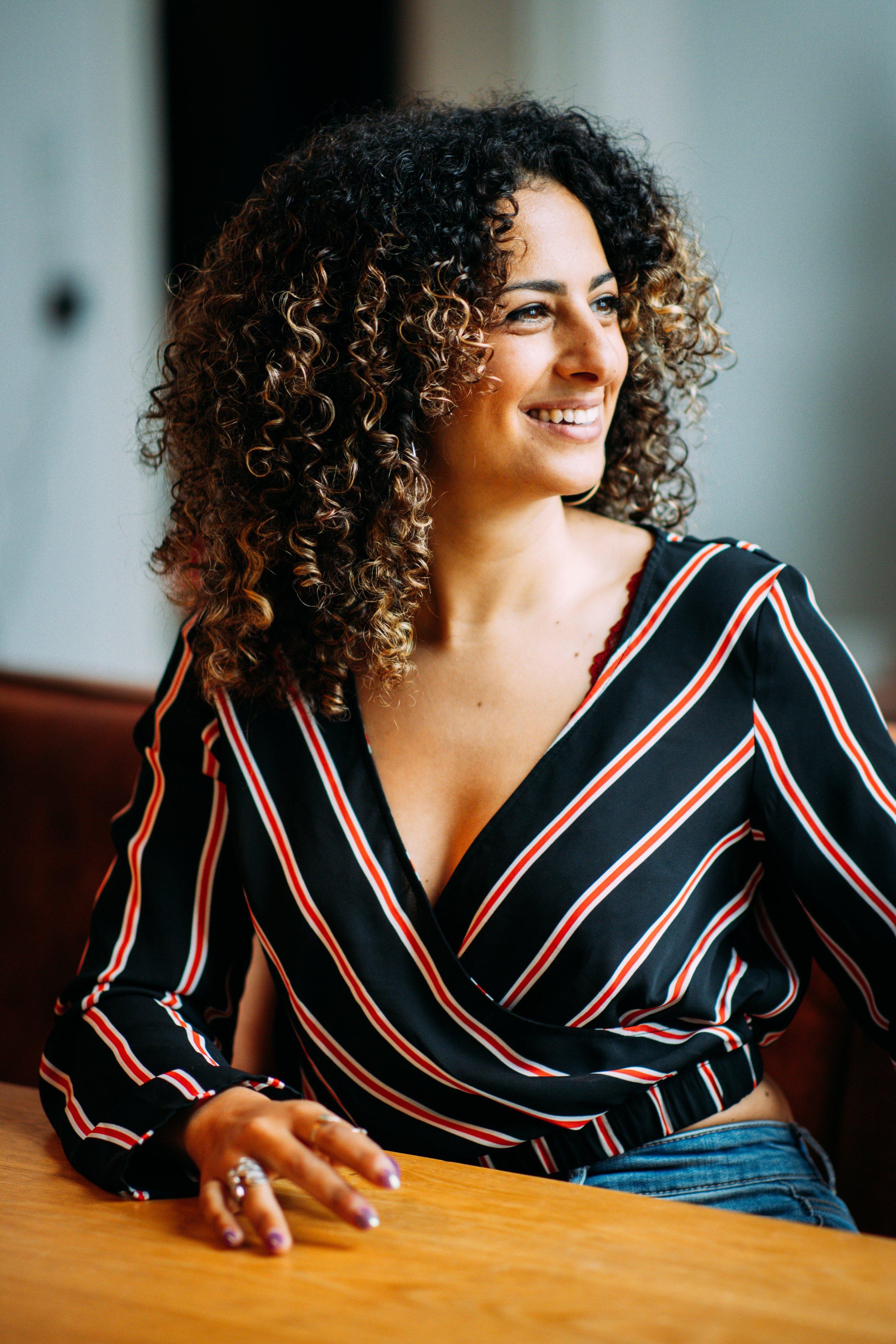 A woman with curly hair smiling, sitting at a wooden table indoors.