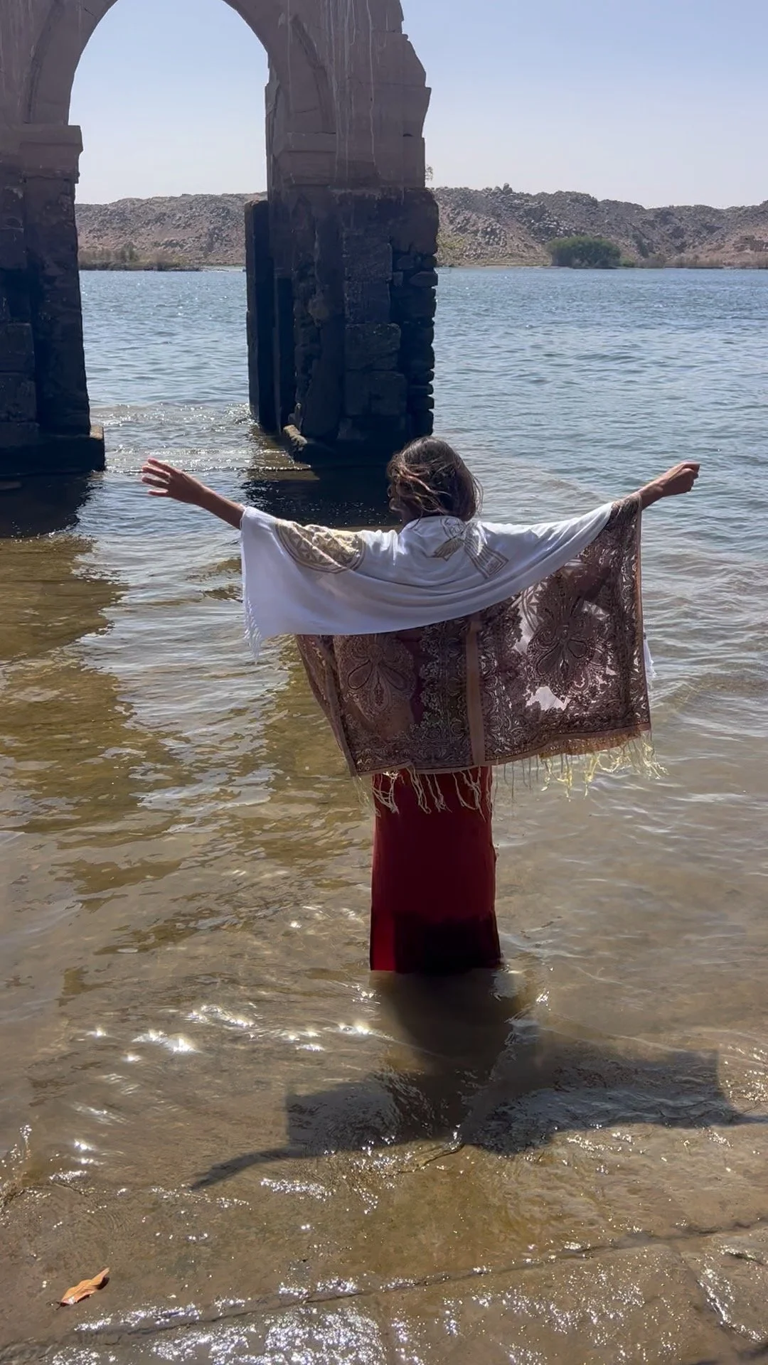 A person standing in shallow water with arms outstretched, wearing a white scarf and a long red dress, near a stone bridge with a river and hills in the background.