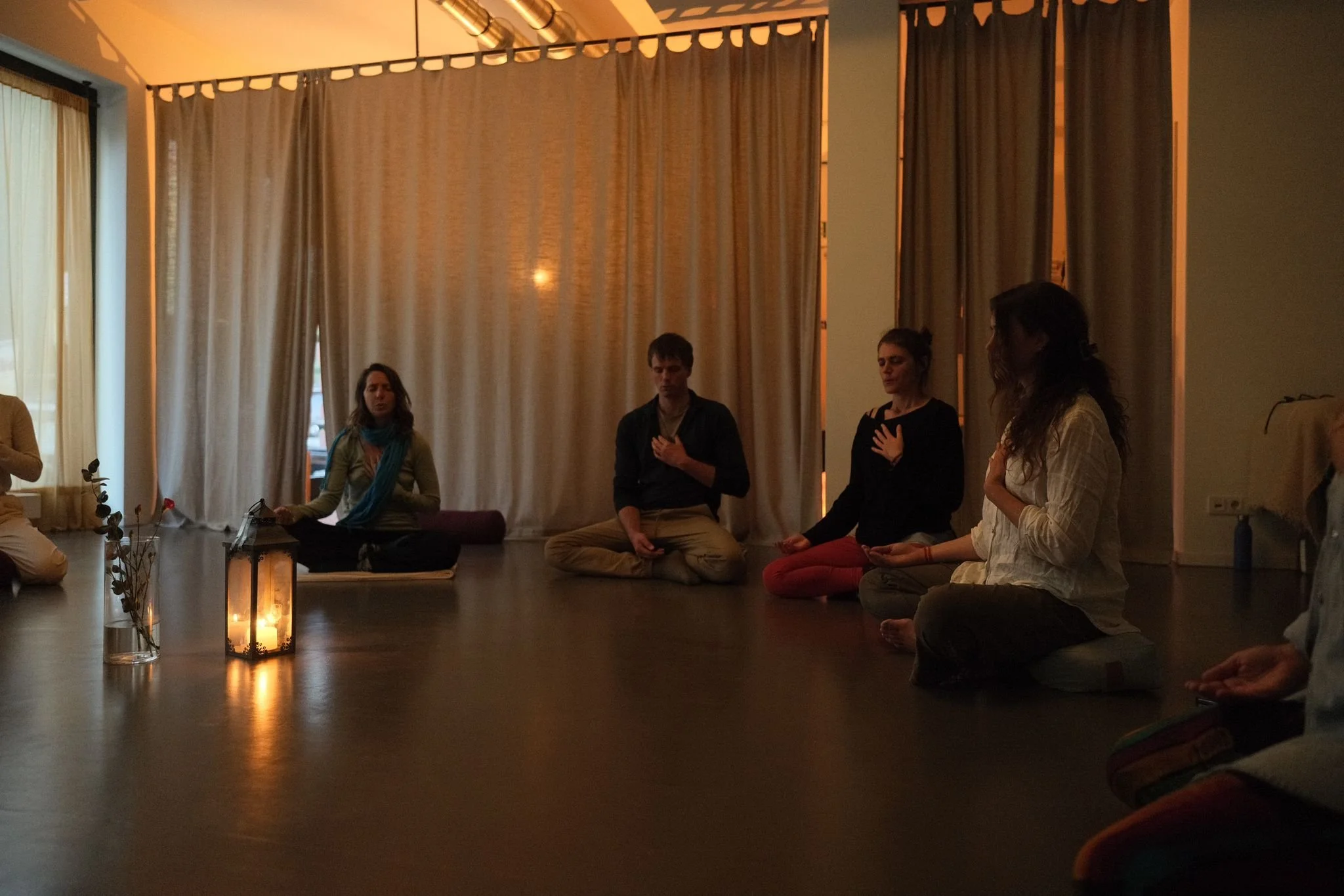 Group of people practicing meditation or mindfulness in a dimly lit room with curtains, candles, and natural light.