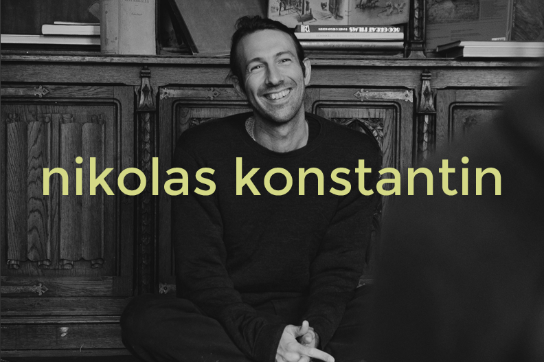 A smiling man sitting on the floor in front of a wooden bookshelf, with the name 'nikolas konstantin' overlaid in yellow text.
