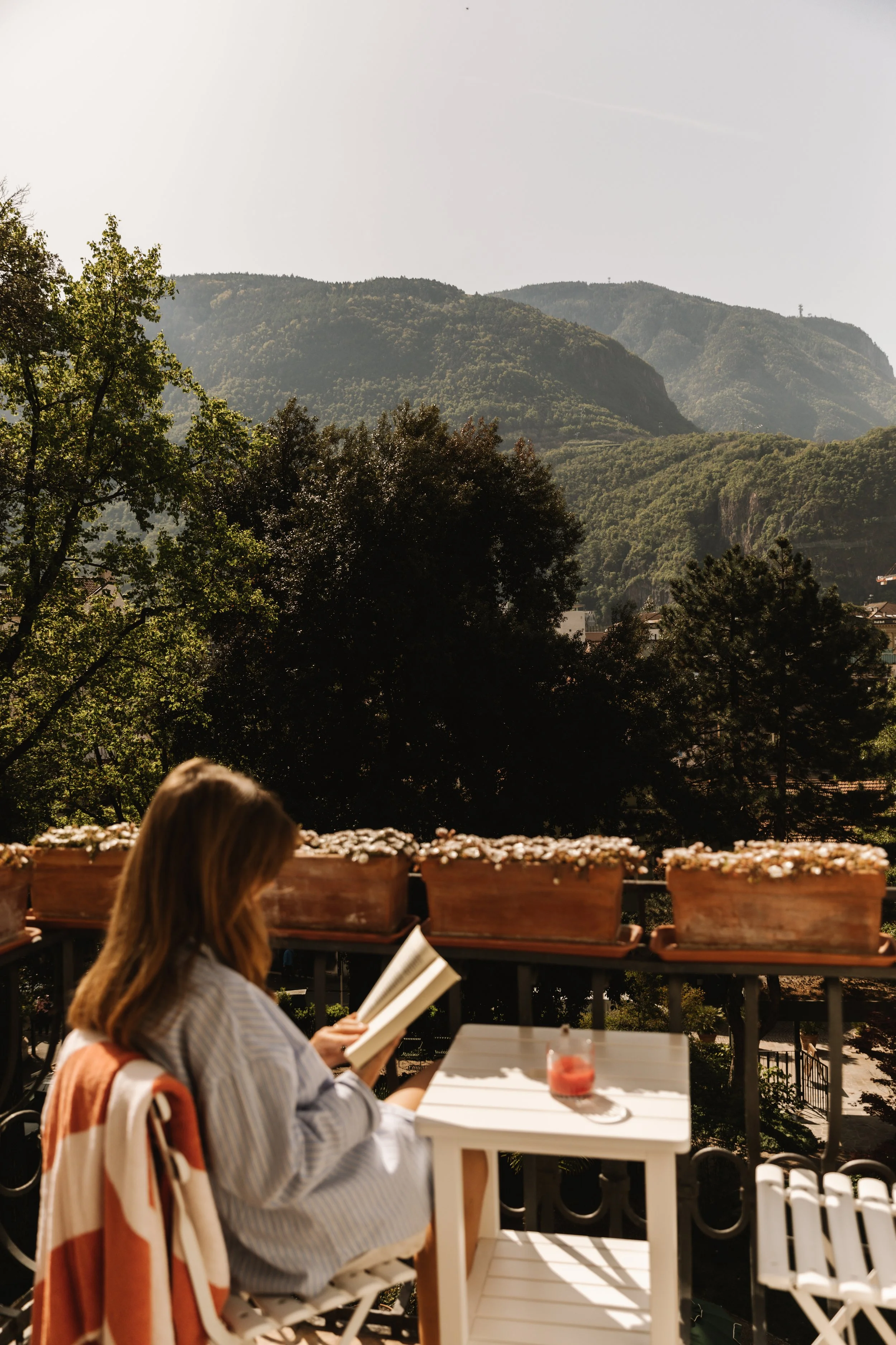 A woman sitting on a balcony, reading a book, with a scenic view of green mountains in the background, potted plants on the railing, and a white table with a drink.
