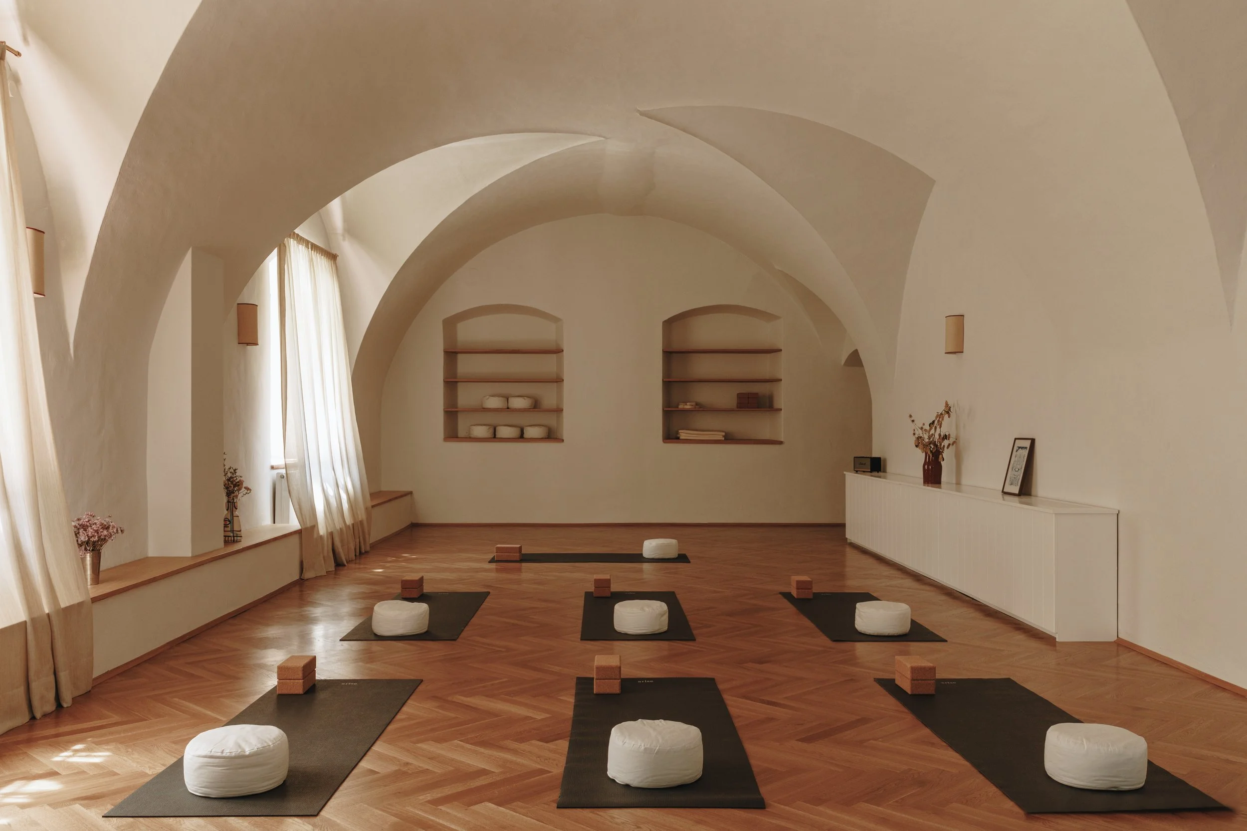 An empty yoga or meditation studio with wooden floors, black mats and white cushions, set up for a class in a bright room with arched ceilings and windows with cream curtains.