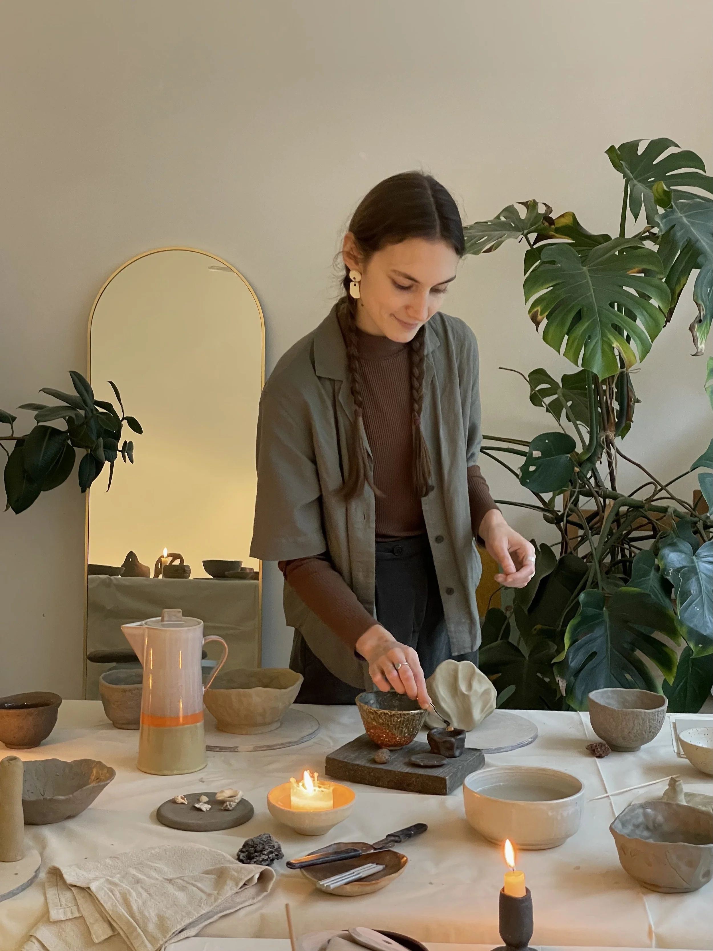 A woman with brown hair in braids looks down while working at a table with pottery and candles, surrounded by green plants.