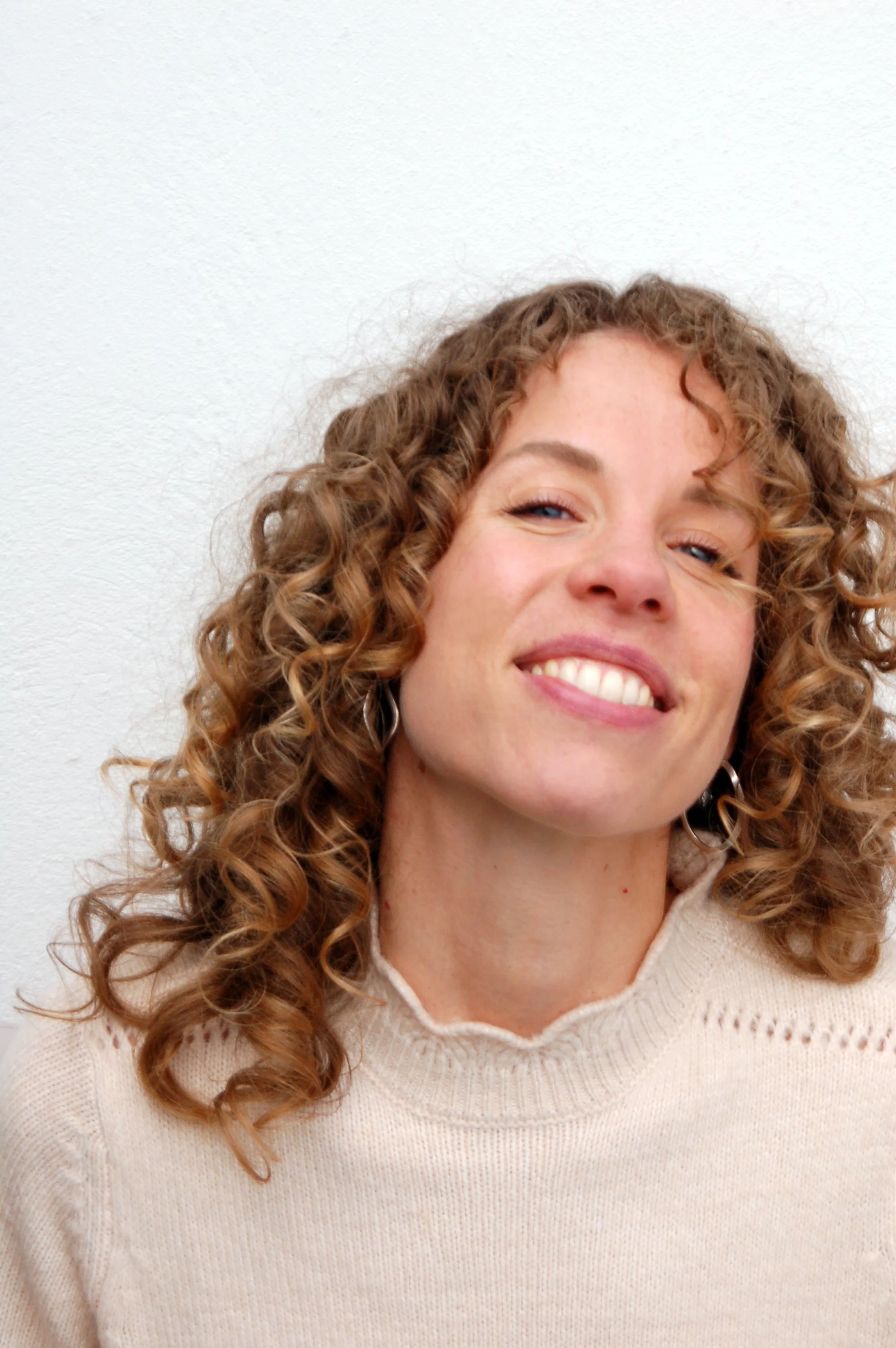 A woman with curly hair smiling and showing her teeth, wearing a beige sweater, standing against a plain white wall.