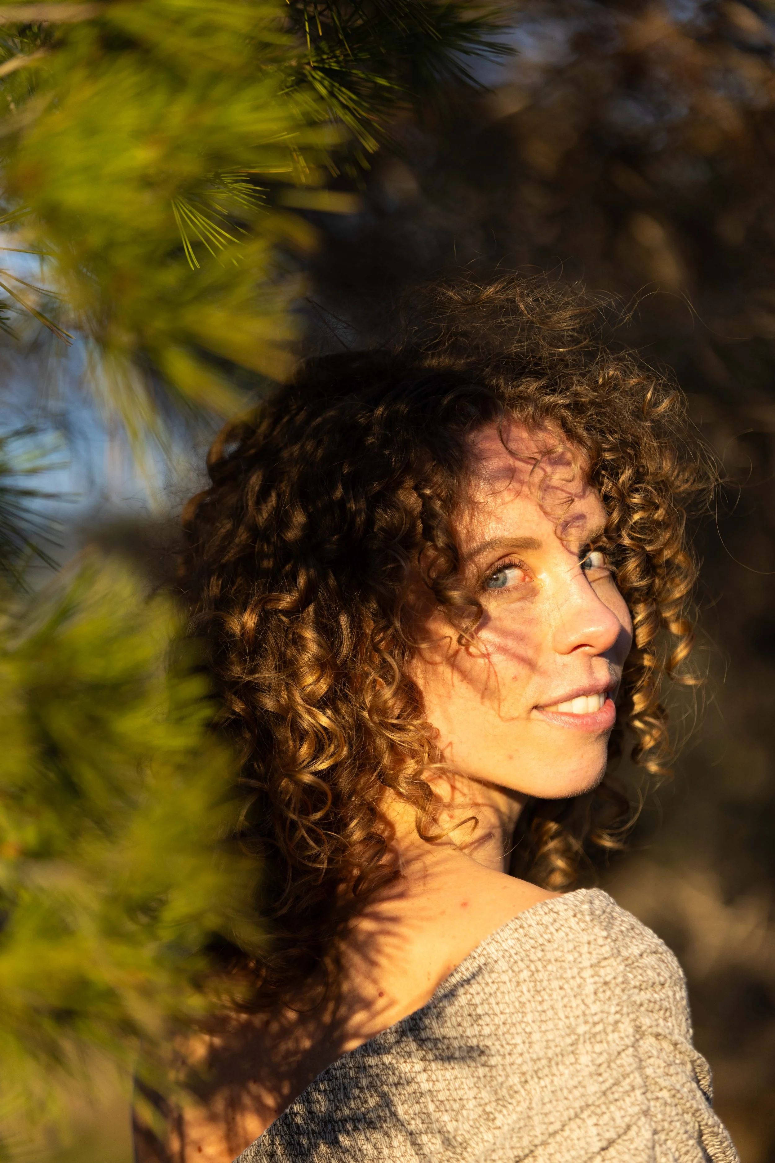 A woman with curly hair turns her head to the side, smiling, as sunlight illuminates her face. She is outdoors near green pine needles.