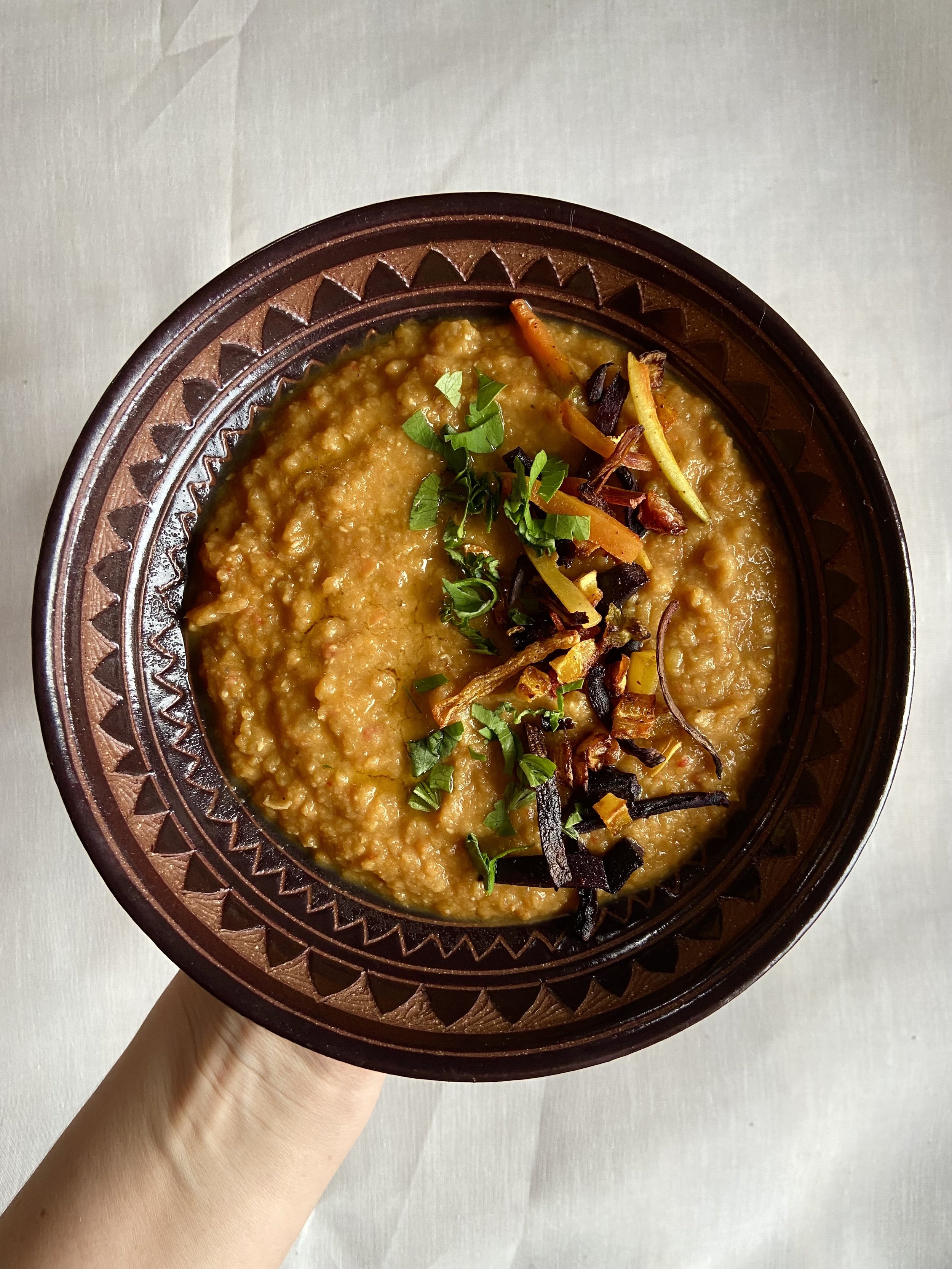 A bowl of Indian lentil soup garnished with chopped herbs and crispy fried vegetables in a decorative brown bowl.