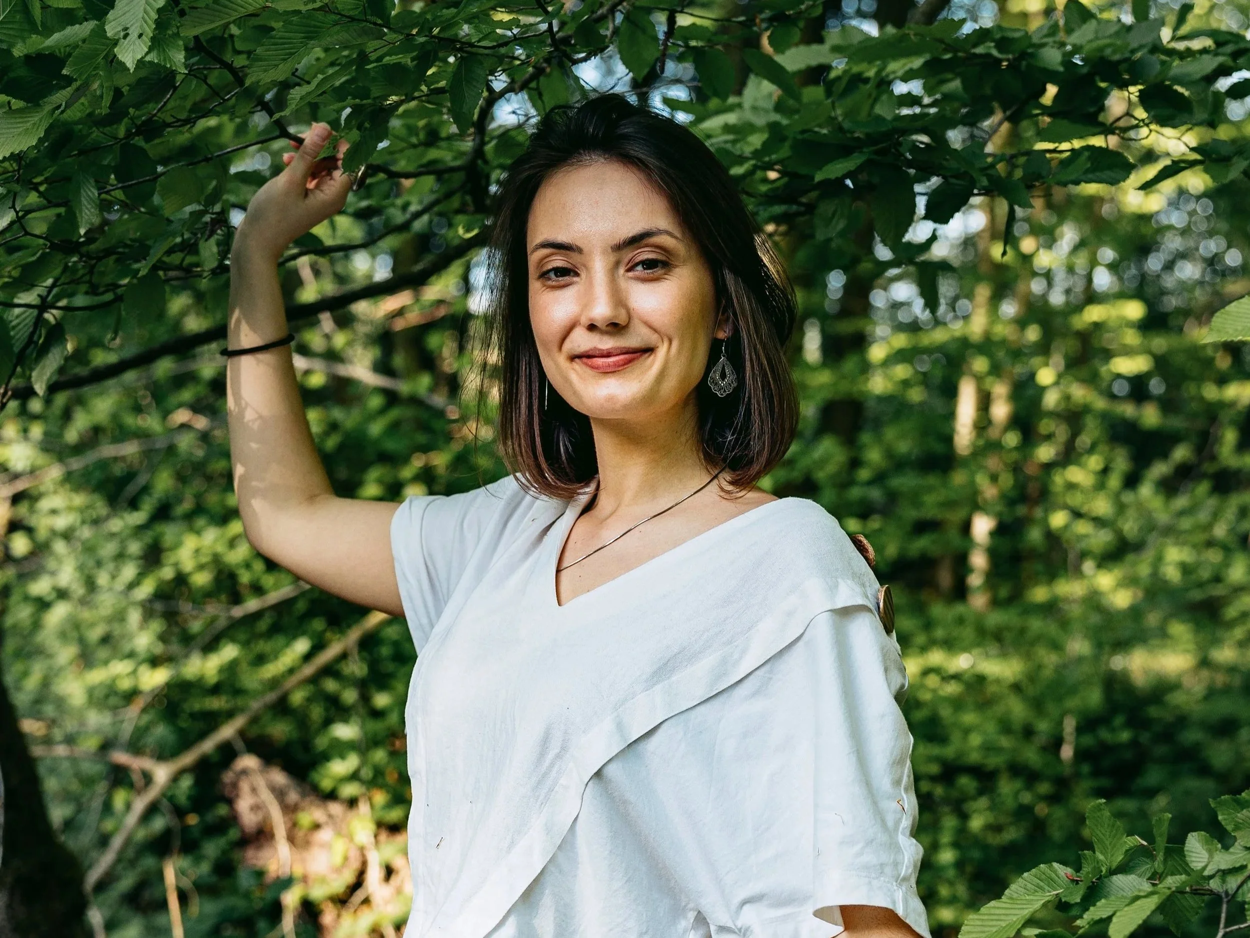 A young woman with shoulder-length dark hair smiling, wearing a white top and earrings, standing in a lush green forest, holding a branch overhead.