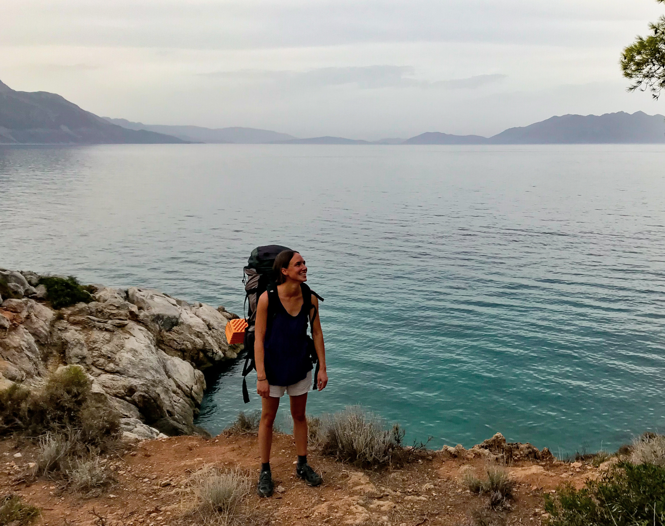 A woman with long hair, wearing a dark tank top, shorts, and hiking boots, stands on a rocky shoreline with a large backpack, smiling and looking to her right, with a calm body of water, distant mountains, and cloudy sky in the background.