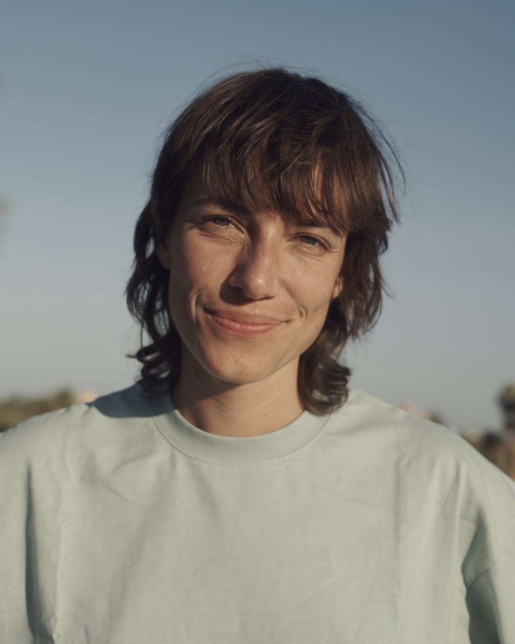 A woman with short brown hair smiling outdoors in sunlight.