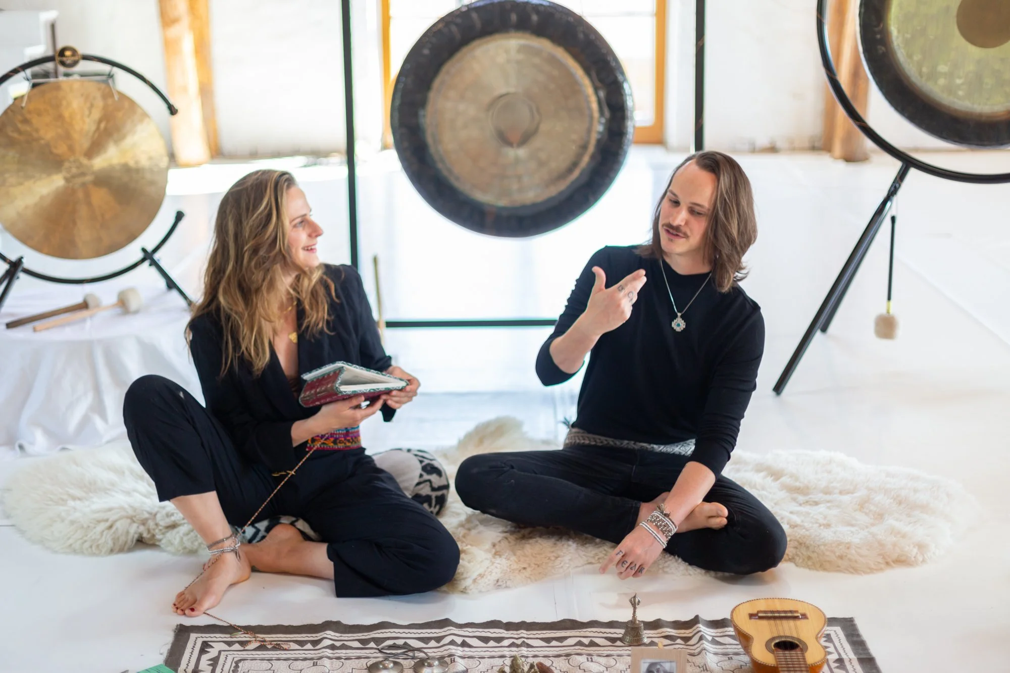 Two people sitting on a fluffy white rug, surrounded by large gongs and musical instruments, engaging in a conversation in a bright room with natural light.