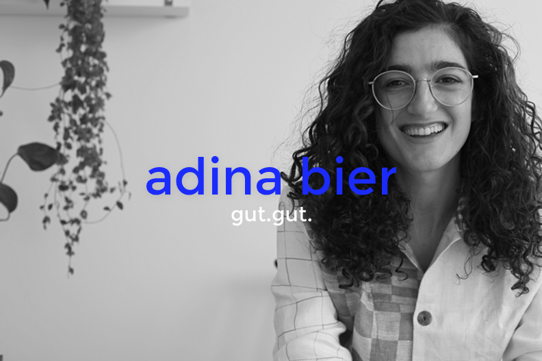 A woman with curly hair and glasses smiling at the camera in front of a wall with hanging plants, with overlaid text "adina bier gut.gut."