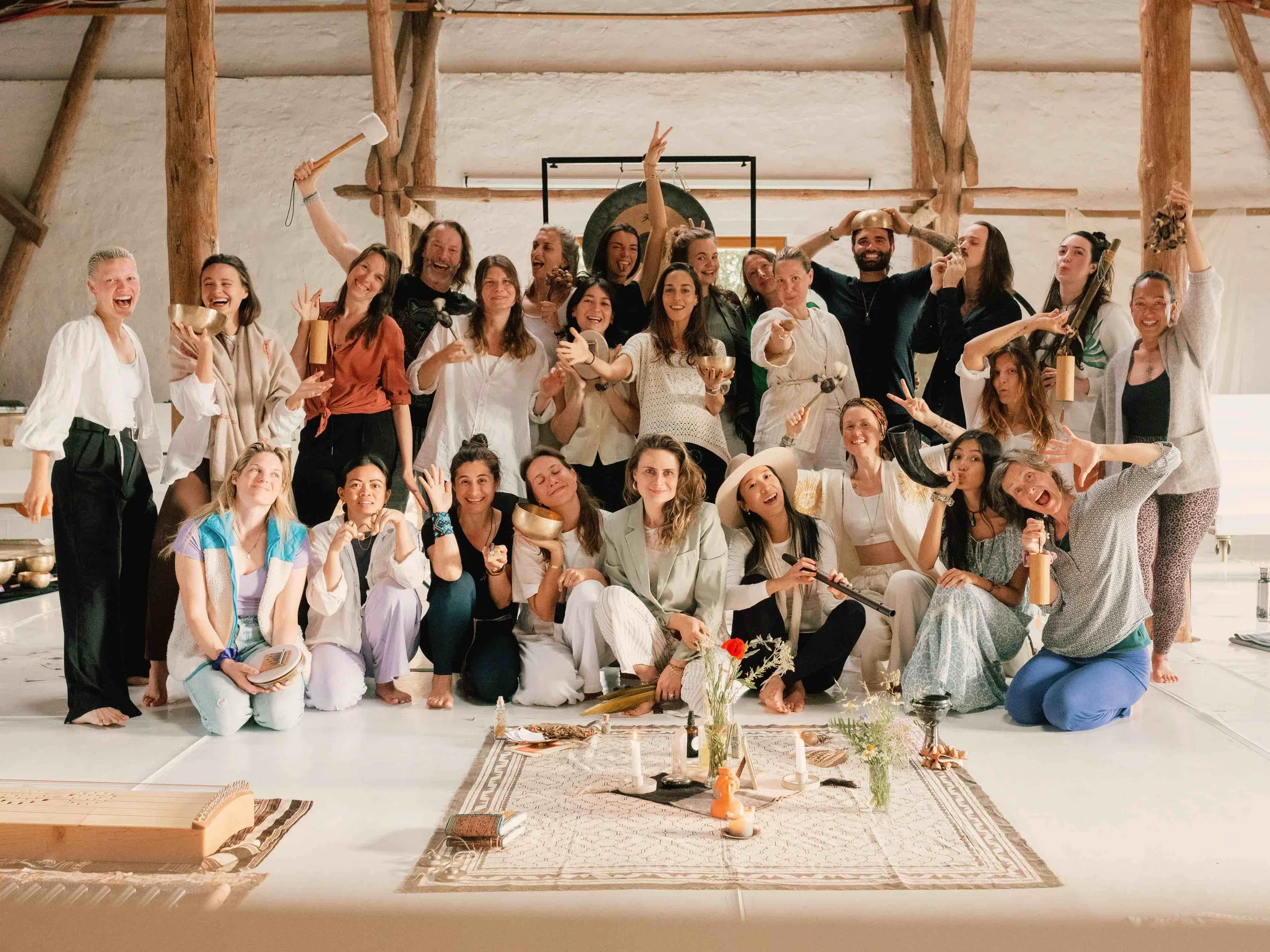 A large group of smiling people gathered in a rustic, wooden-ceiling room, celebrating together with musical instruments, bowls, and a decorated altar on the floor.