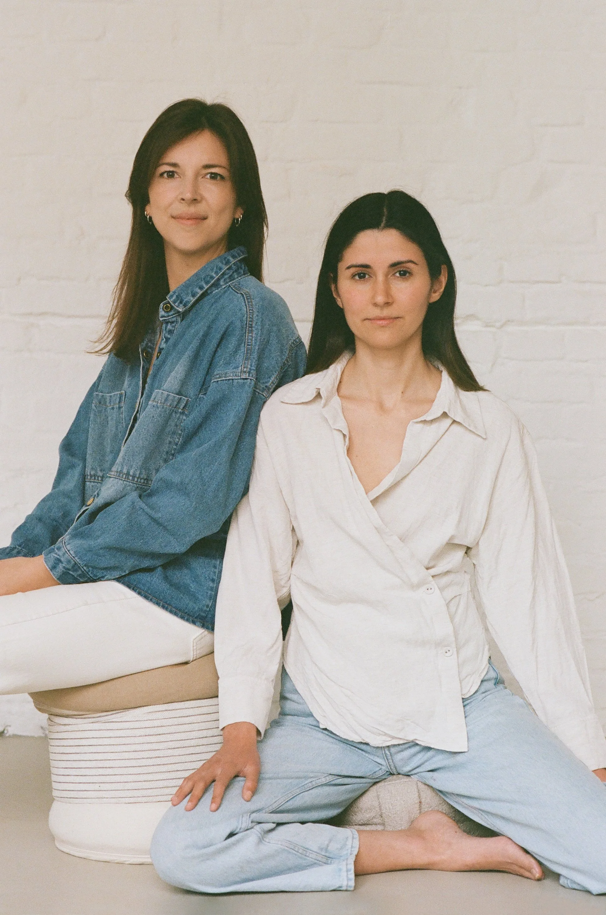 Two women sitting against a white brick wall, one in a denim jacket and the other in a light-colored shirt and jeans.