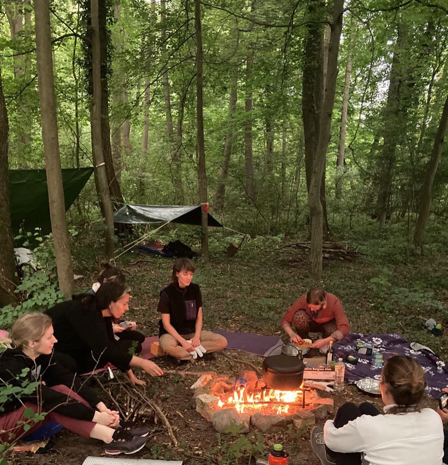 Group of people sitting around a campfire in a forested area with tents and camping gear nearby