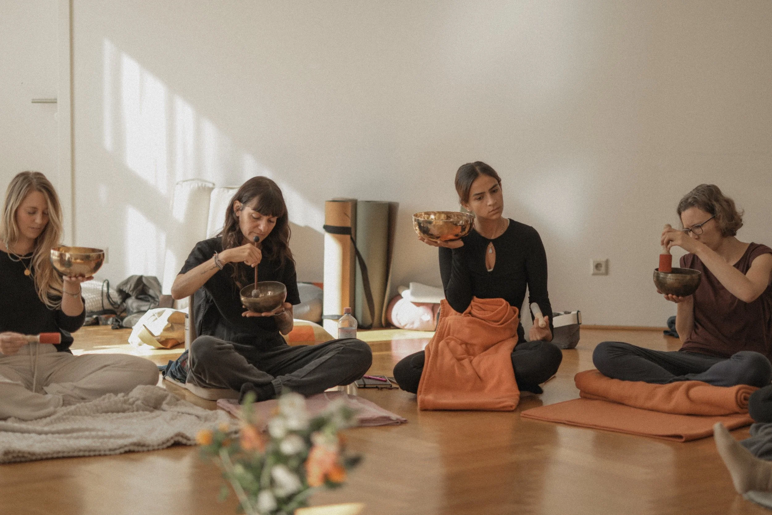 Four women sitting on the floor in a meditation or yoga class, holding singing bowls and other musical instruments in a minimalistic room with wooden floor and white wall.