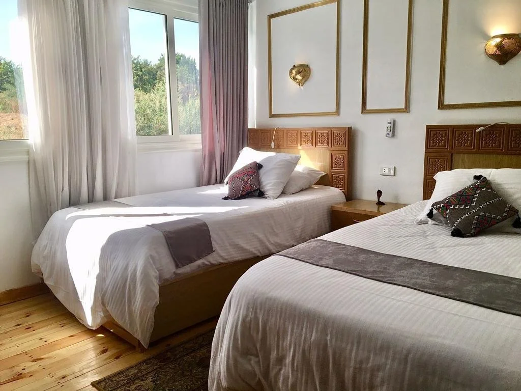A bedroom with two beds, white linens, decorative pillows, a window with curtains, and wall-mounted lights.