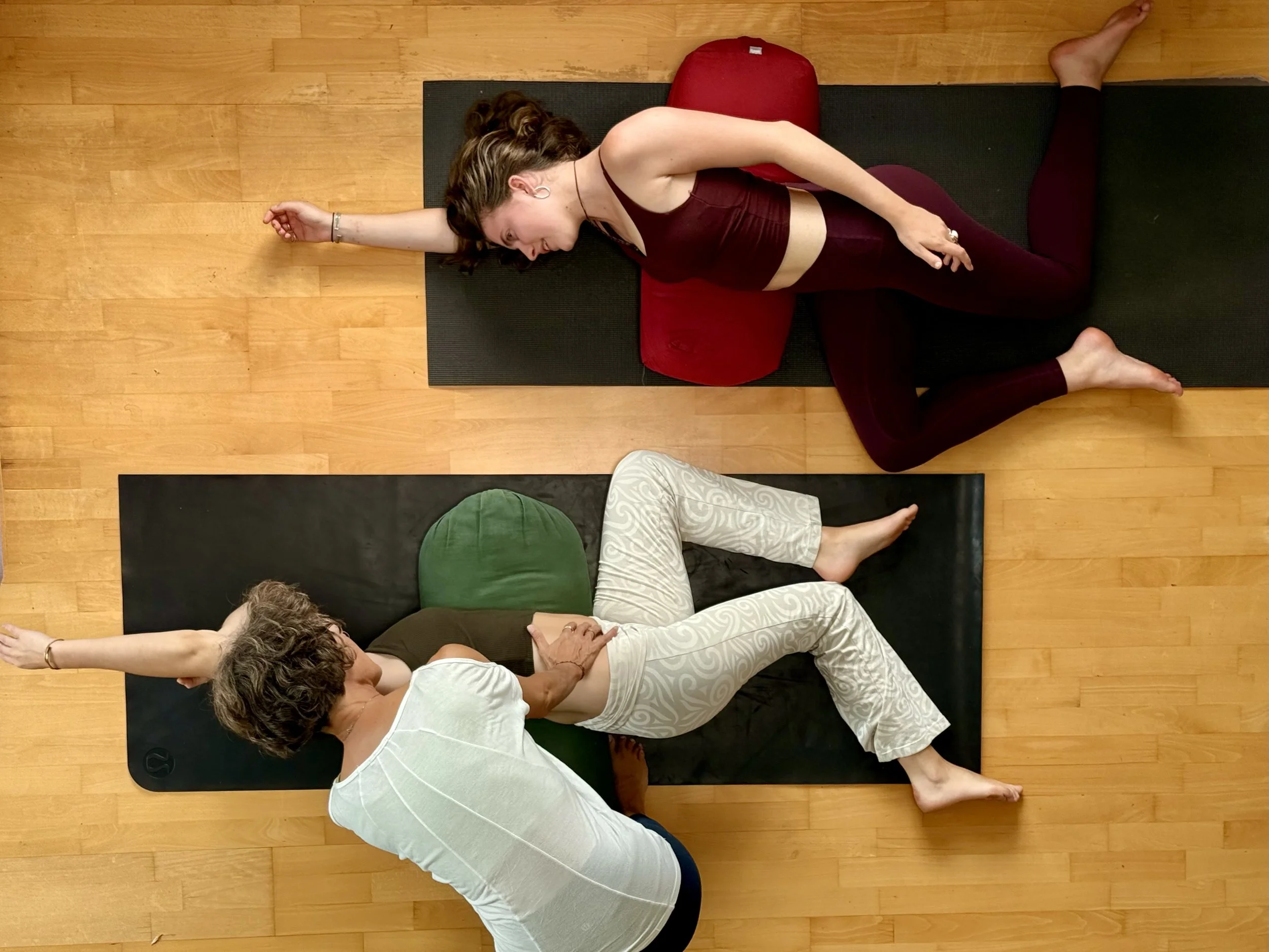 Two women practicing yoga on black mats, with bolsters, in a room with wooden floors. One woman is lying on her side with her arm extended, the other woman is lying on her stomach with her hand on her abdomen.