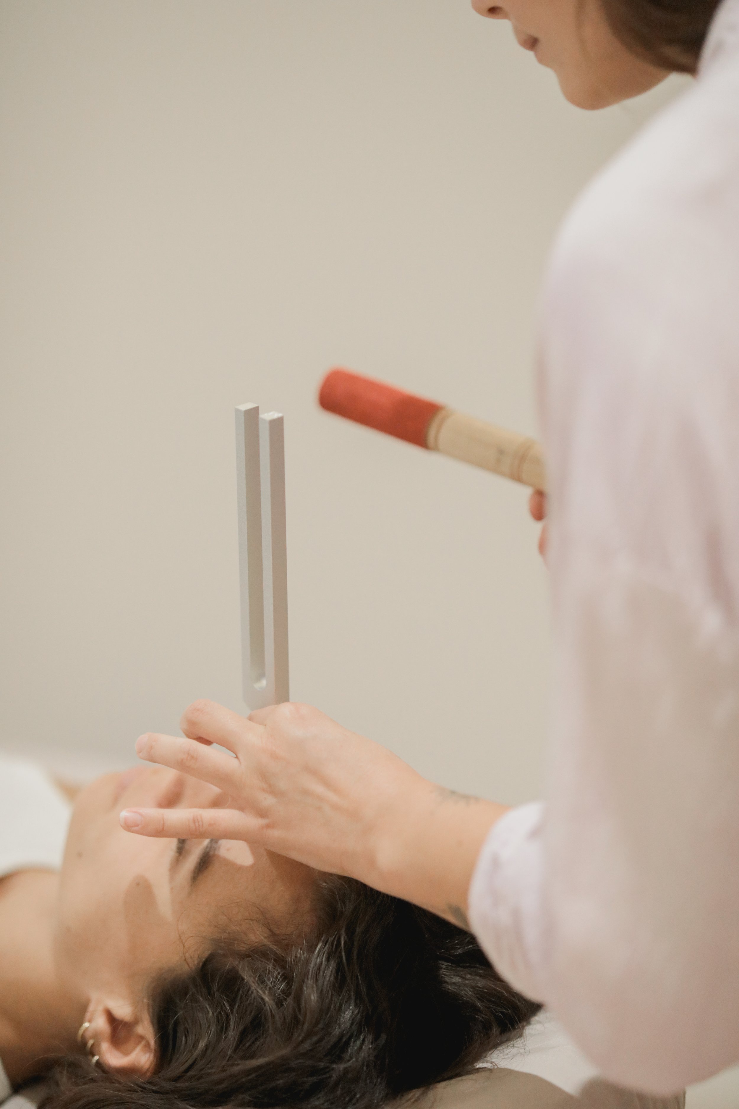 A person with dark hair lying down with closed eyes receives a tuning fork test on their forehead from a practitioner.