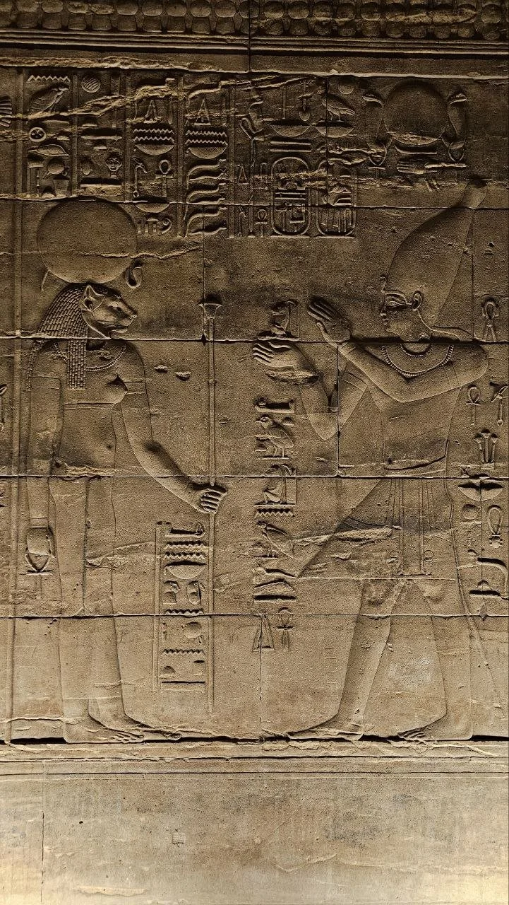 Ancient Egyptian carved stone wall depicting a Pharaoh and a deity exchanging objects, with hieroglyphic inscriptions above them.