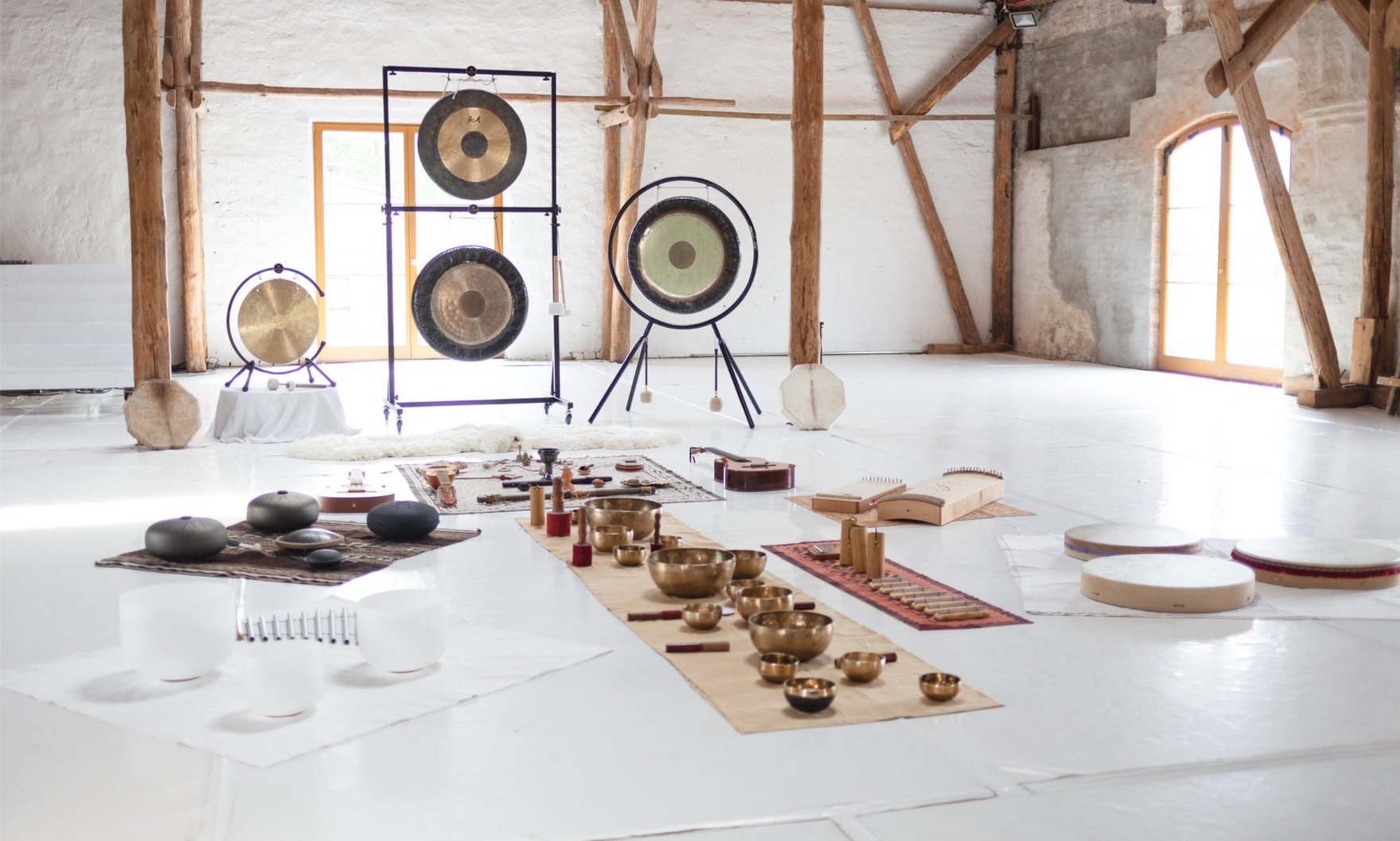 An indoor space with white walls, large windows, and exposed wooden beams, displaying musical and sound instruments including gongs, singing bowls, and wooden percussion instruments arranged on the floor.