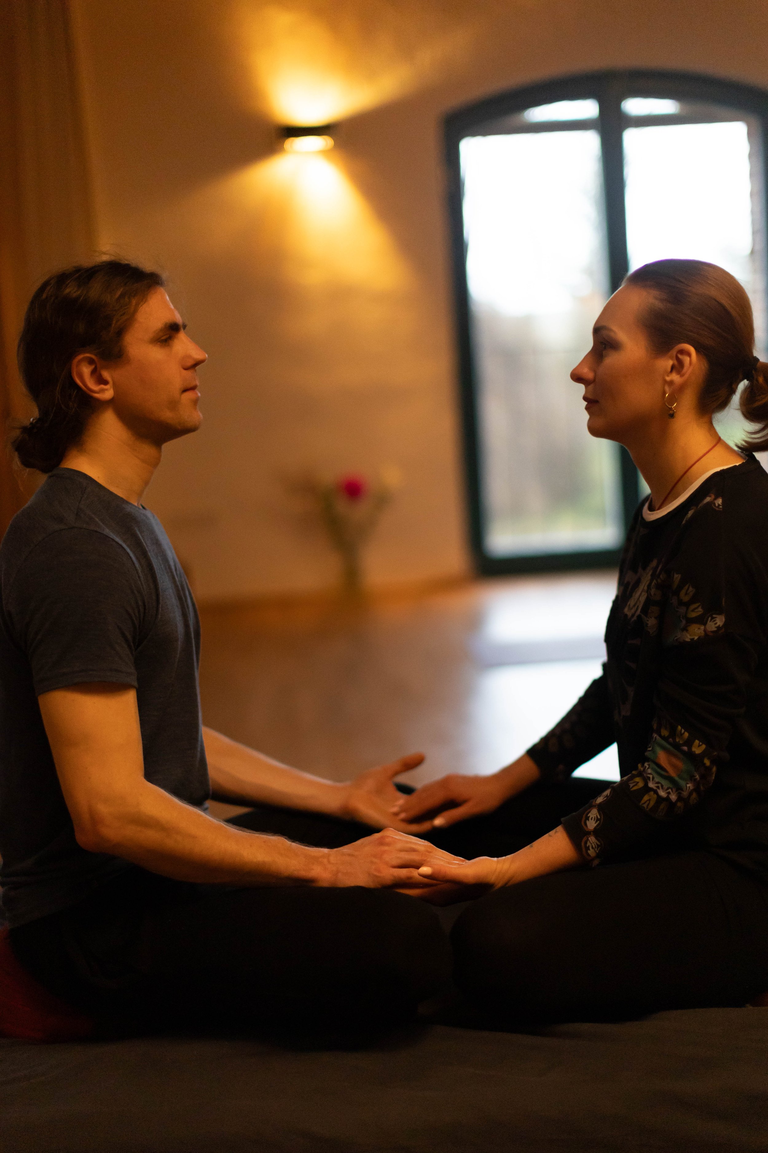 A man and a woman sitting on the floor facing each other, holding hands, in a room with a large window and warm lighting.