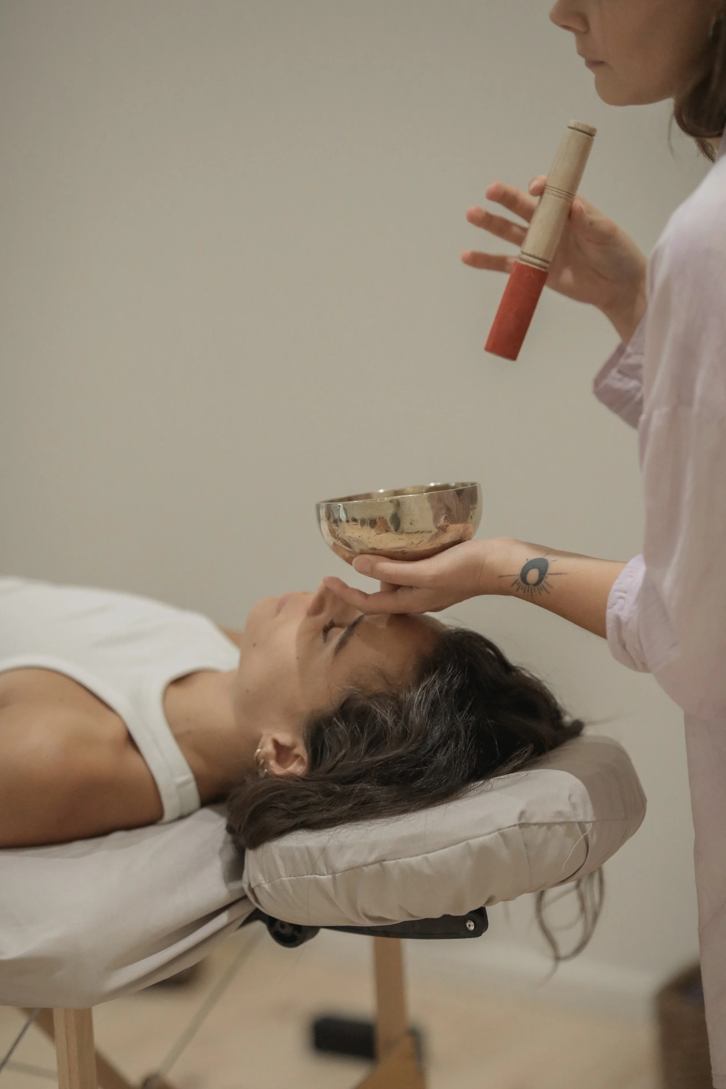 A woman lying on a massage table with a pillow under her head, receiving a spiritual or energy healing from a practitioner holding a Tibetan singing bowl, with the practitioner using a mallet. The practitioner is standing next to the woman, who has a