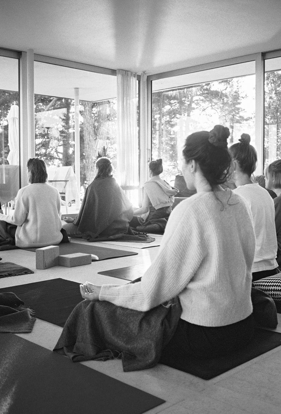 Group of people meditating in a spacious, bright room with large windows showing trees outside, seated on mats with some in traditional robes and others in casual clothes.